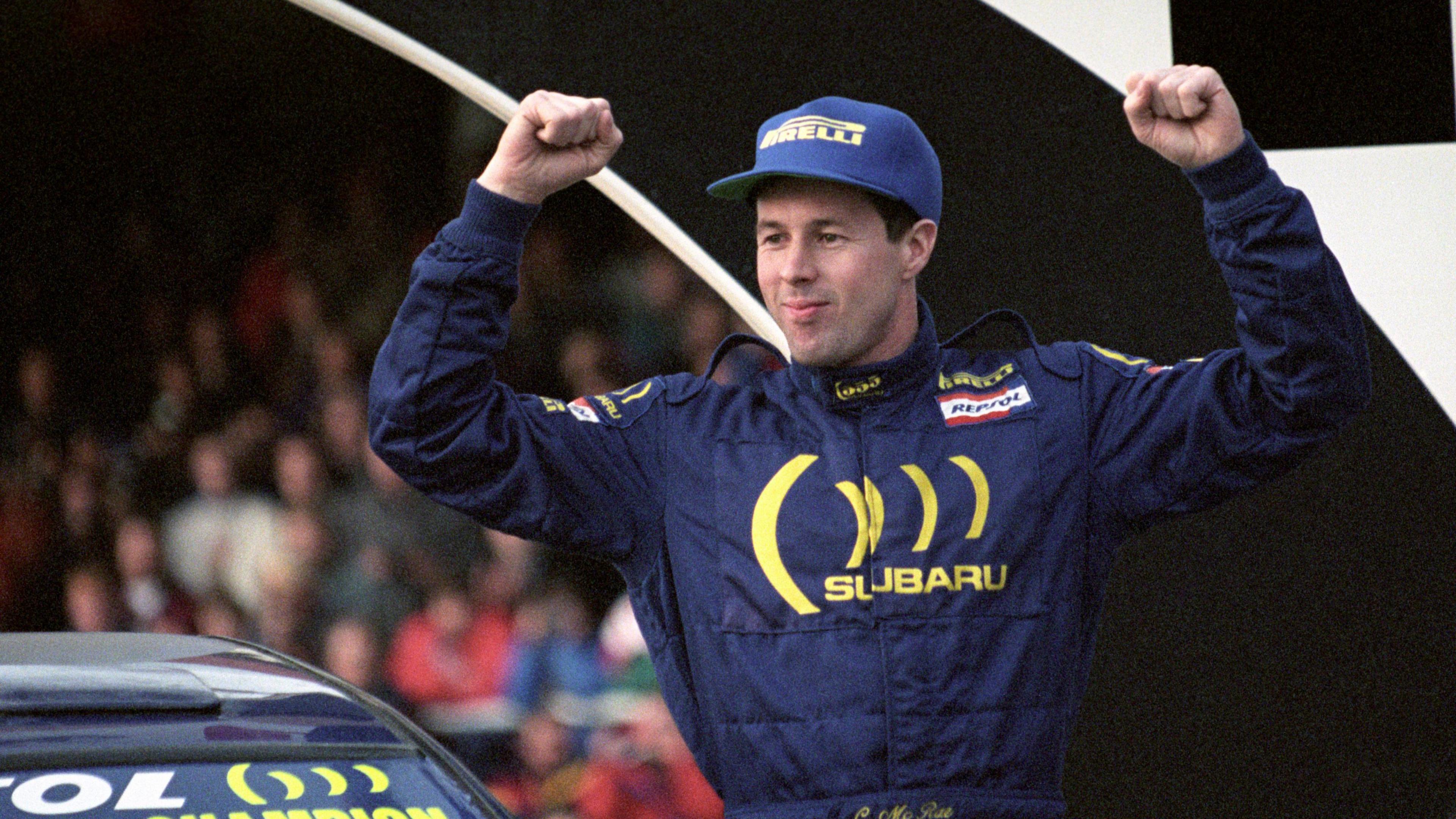 Colin McRae in a blue boiler suit with Subaru branding. He also has a blue Pirelli baseball cap. His arms are in the air in celebration. He's standing next to a rally car, in front of a crowd of people