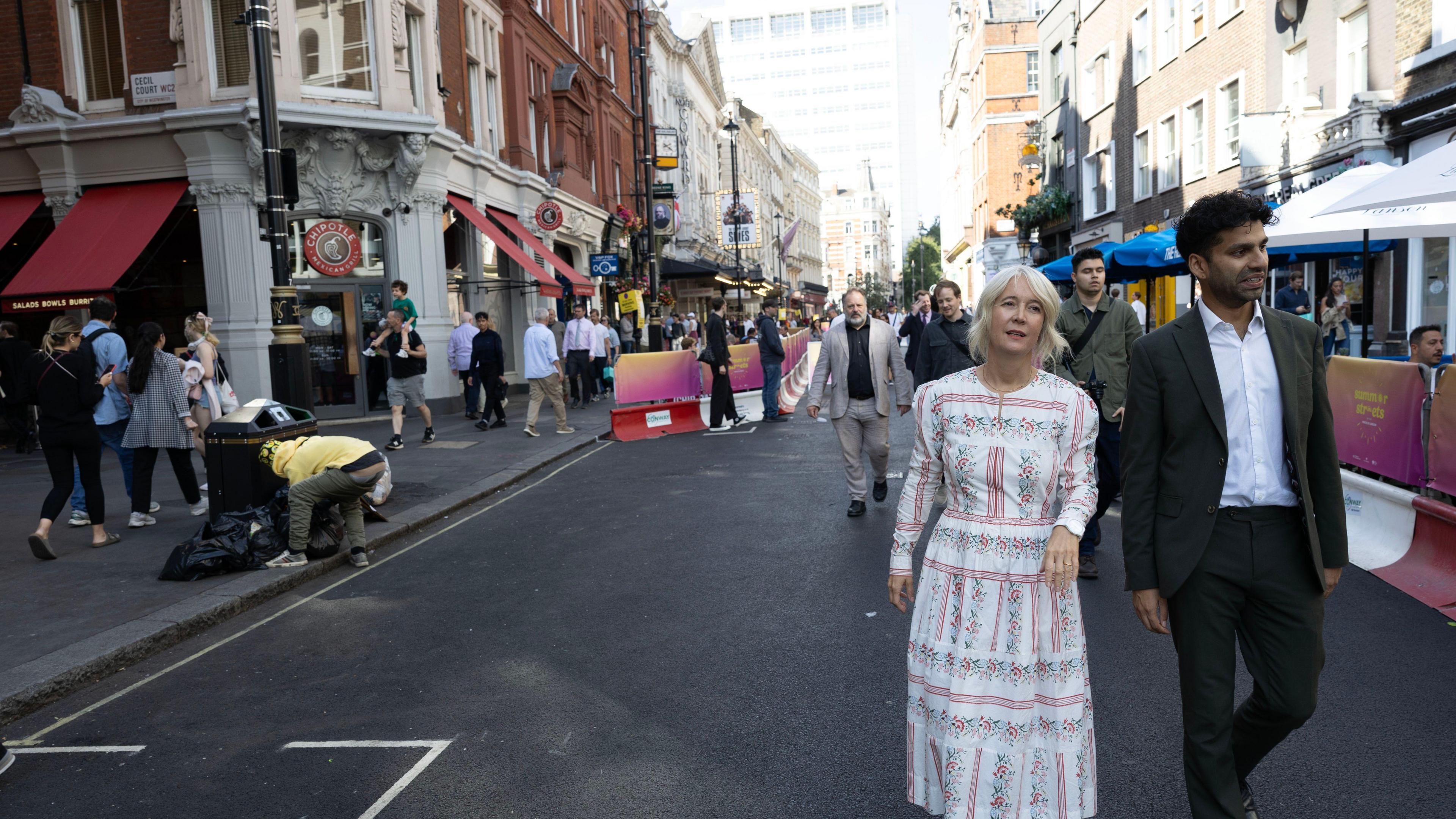 Justine Simmons is seen in a patterned white dress and yellow shoes walks alongside a man in a dark suit on a busy city street, with people and shops visible in the background.