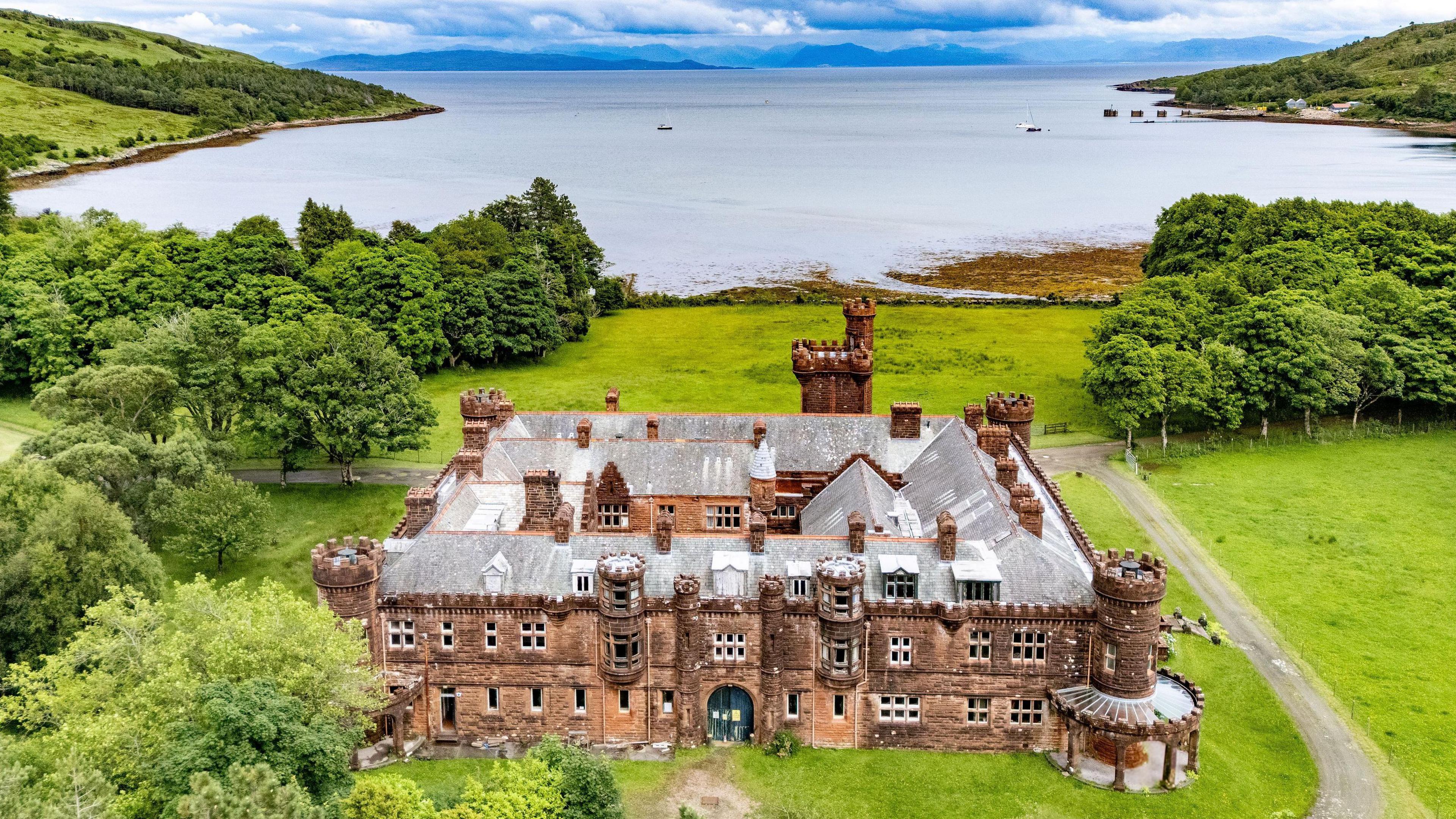 An aerial view of the red sandstone lodge with its ornate turrets and battlement-like rooftops. The lodge is set in an grounds with large areas of grass and trees. In the background is the sea, with a few boats dotted around a bay, and mountains in the distance.