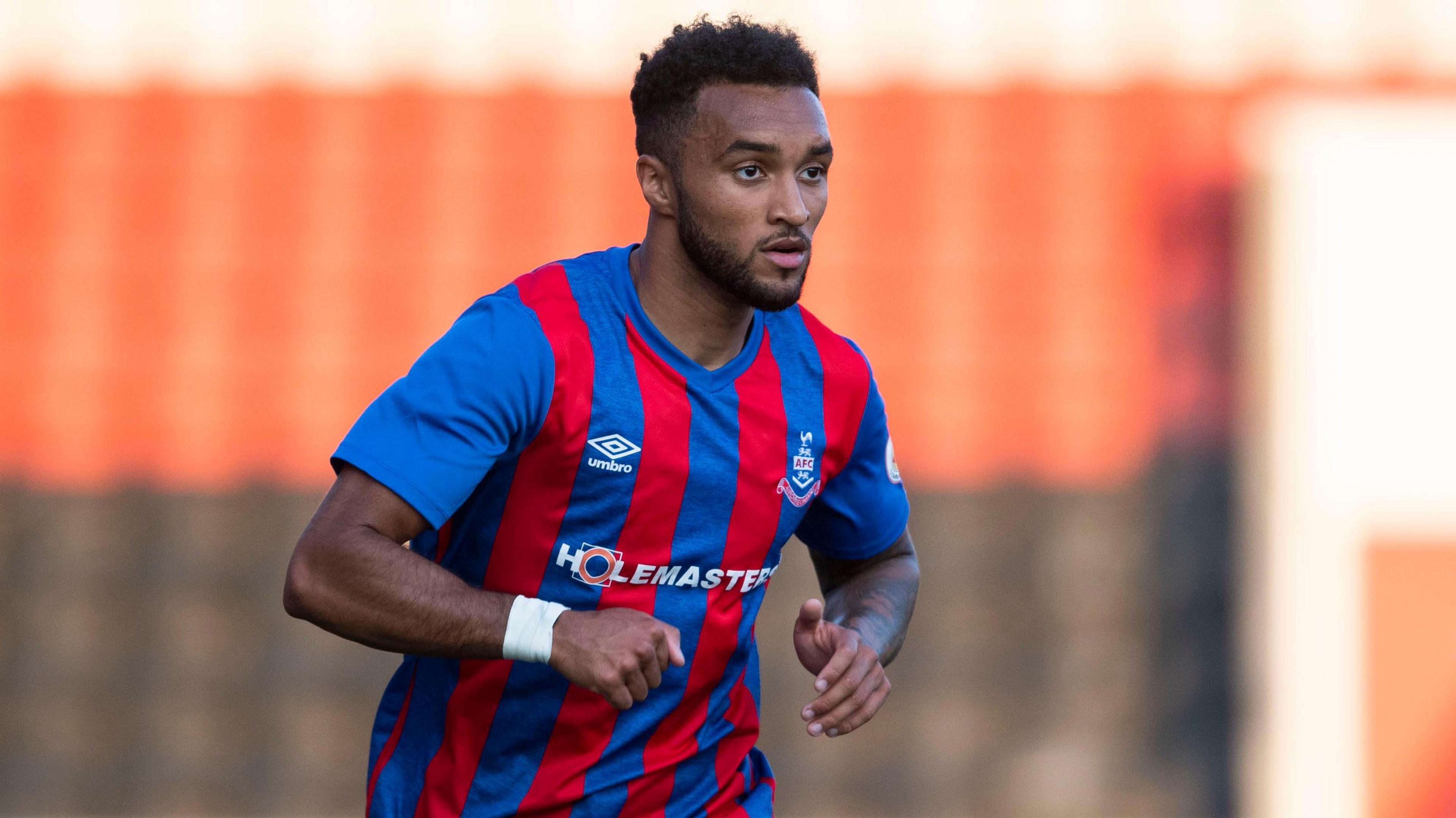 A footballer in a red and blue striped kit has his eyes fixed on the ball