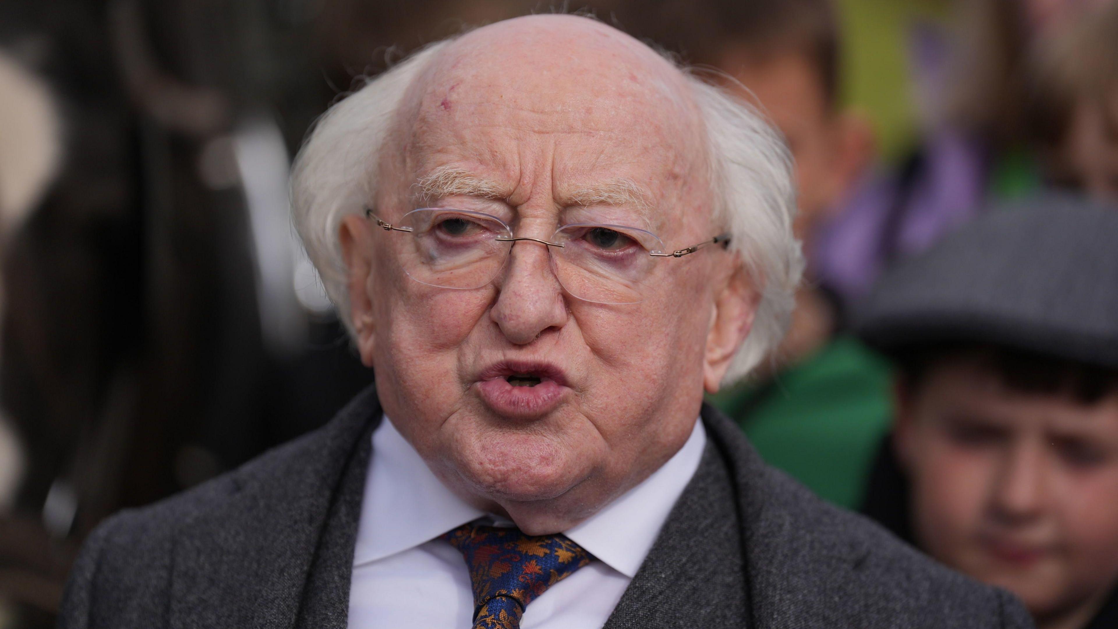 A close-up image of Michael D Higgins, who is attending the Ploughing Championships in County Offaly. President Higgins is an older man, who is balding with long white hair. He has frameless, rectangle glasses and is mid-speech. He is wearing a grey, woolen suit wth a white suit and blue and orange patterned tie.