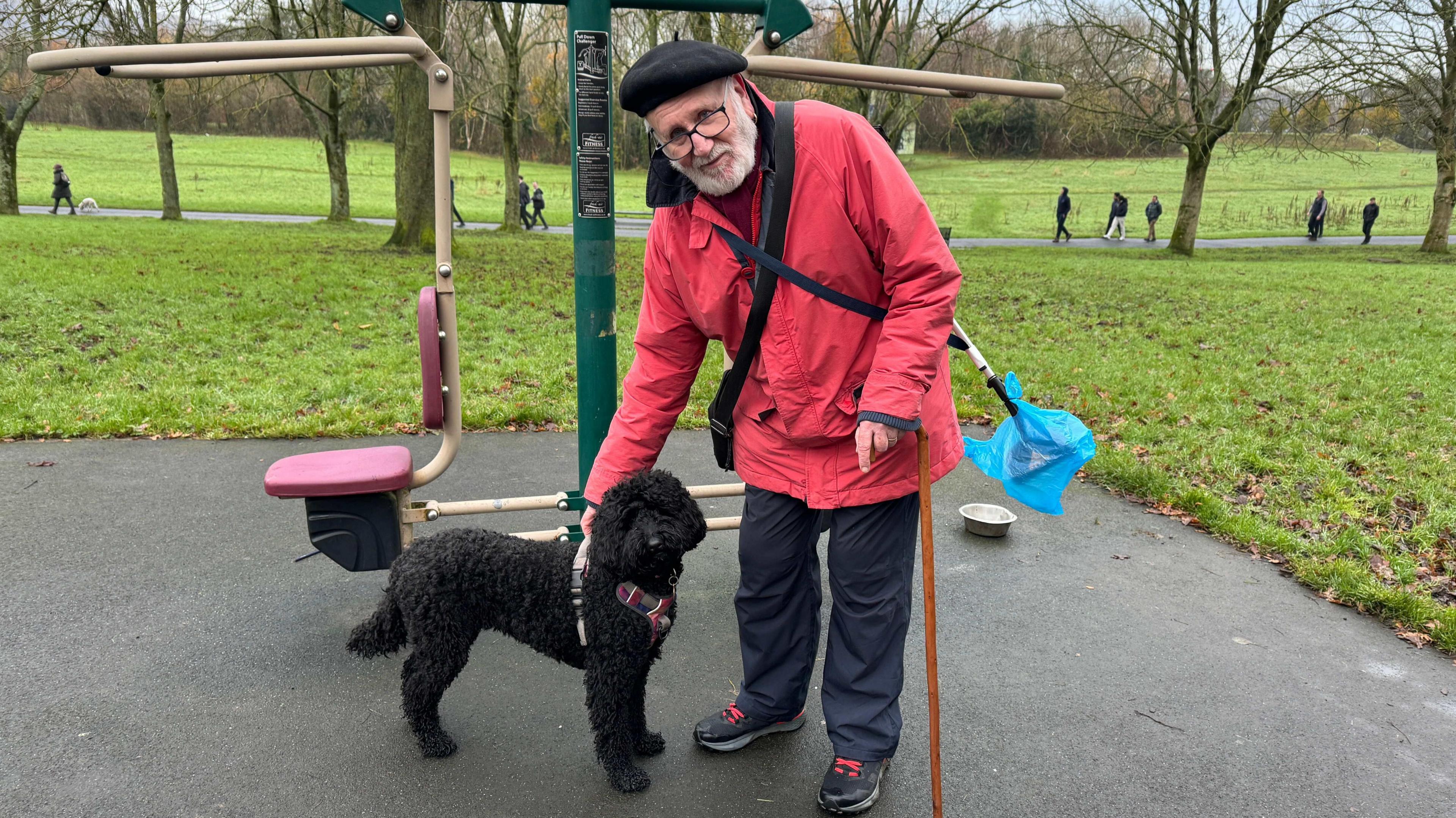 A man wearing a red rain coat pets his black poodle dog in the middle of a park. Around him is grass, some trees in the background, and on a distant path you can see a number of other walkers. 