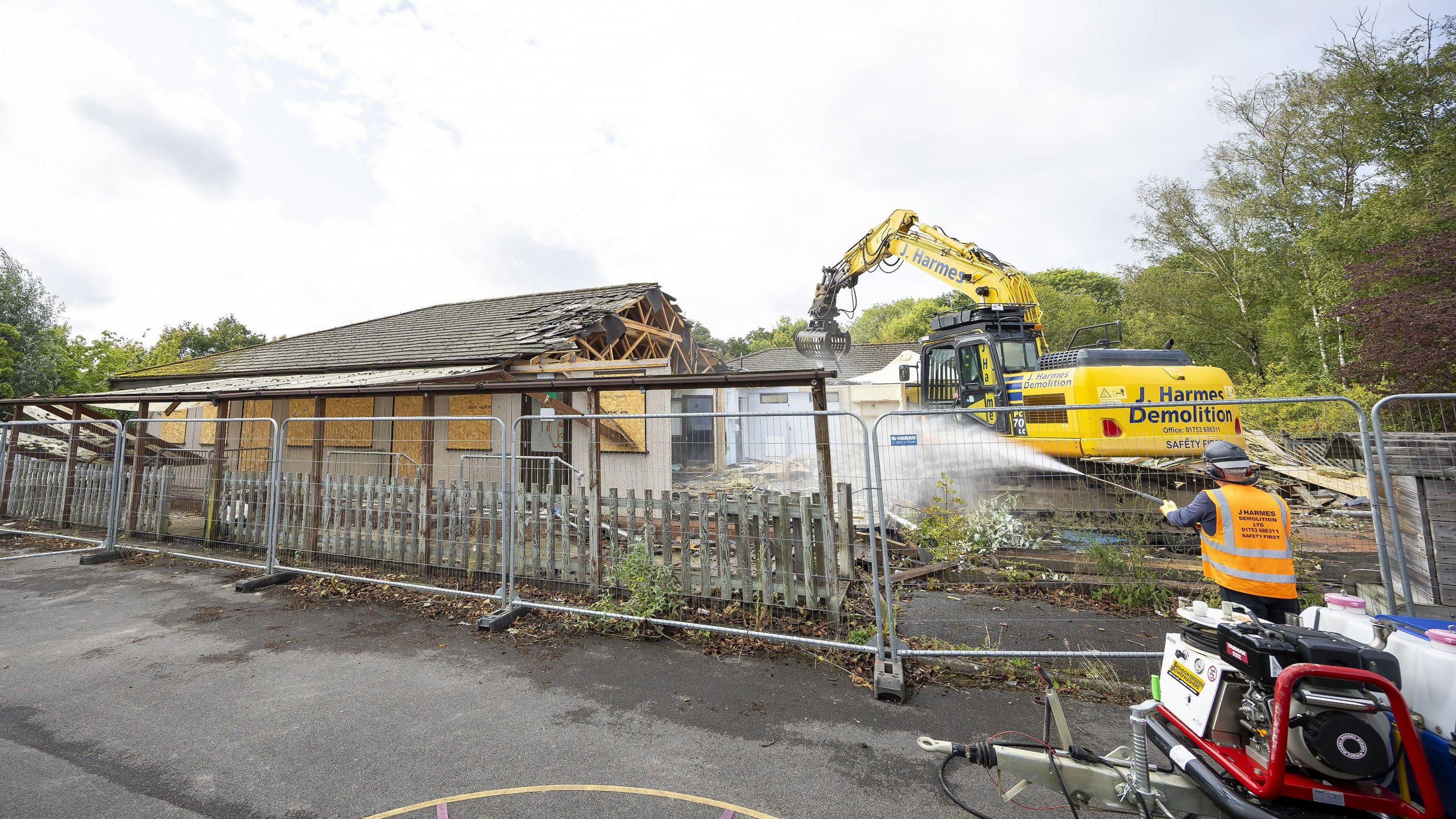 A school building, which has boards in its windows, being demolished by a large piece of machinery as a worker sprays water at it from a hose or a pressure washer.