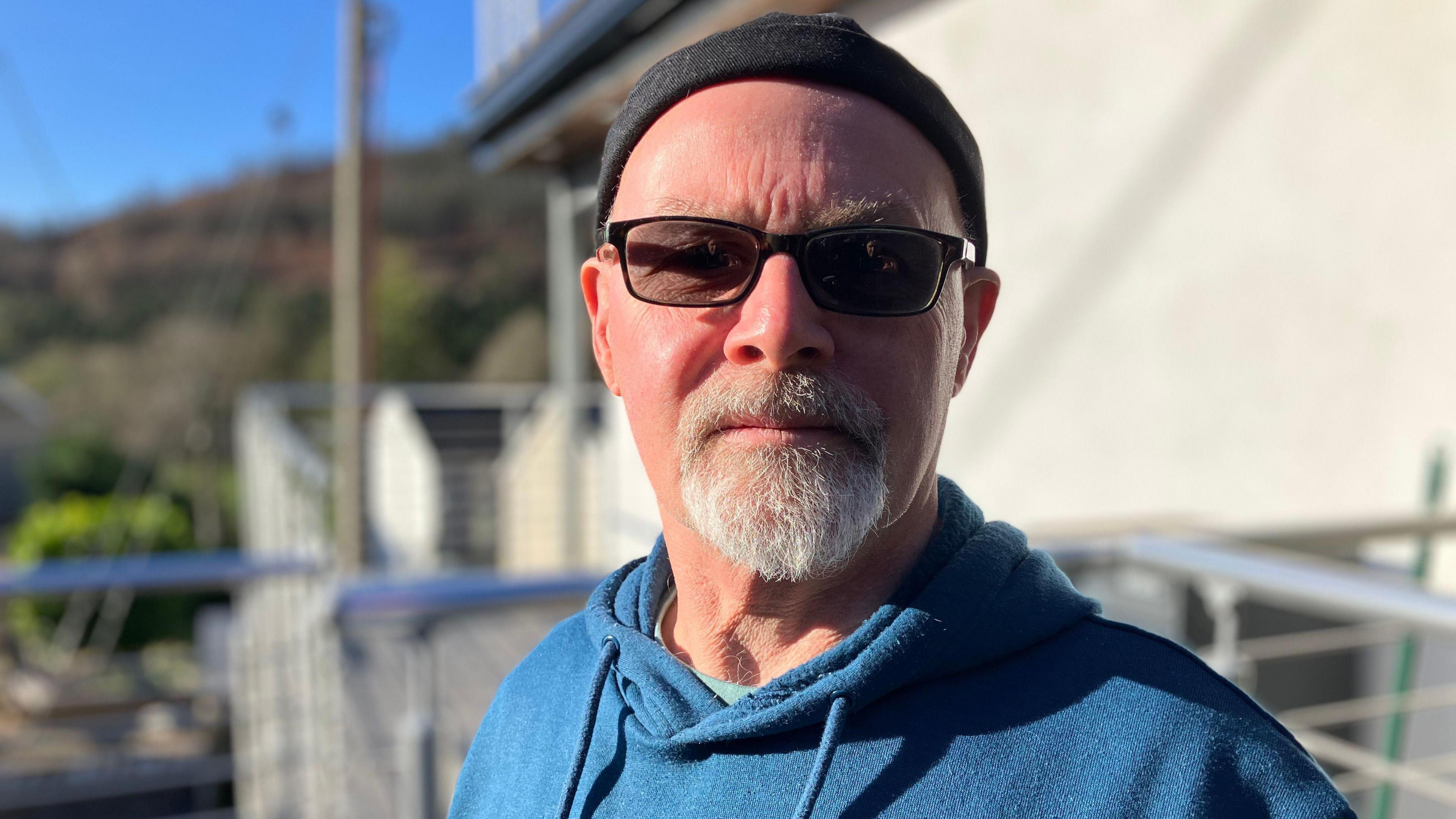 Leslie Morgan is wearing a beanie hat and blue hoodie and is standing on his balcony which overlooks the valley in front of him and the tip behind.