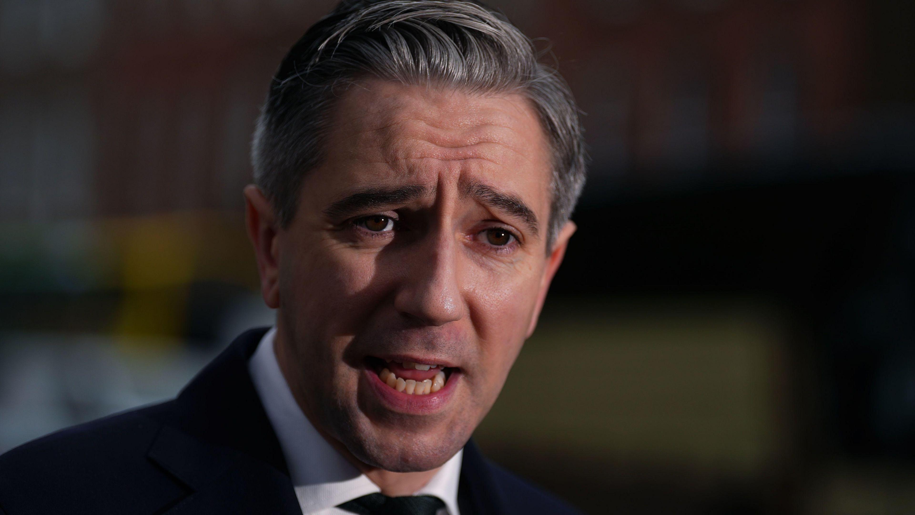 Simon Harris with grey hair wearing a dark suit, white shirt and black tie.