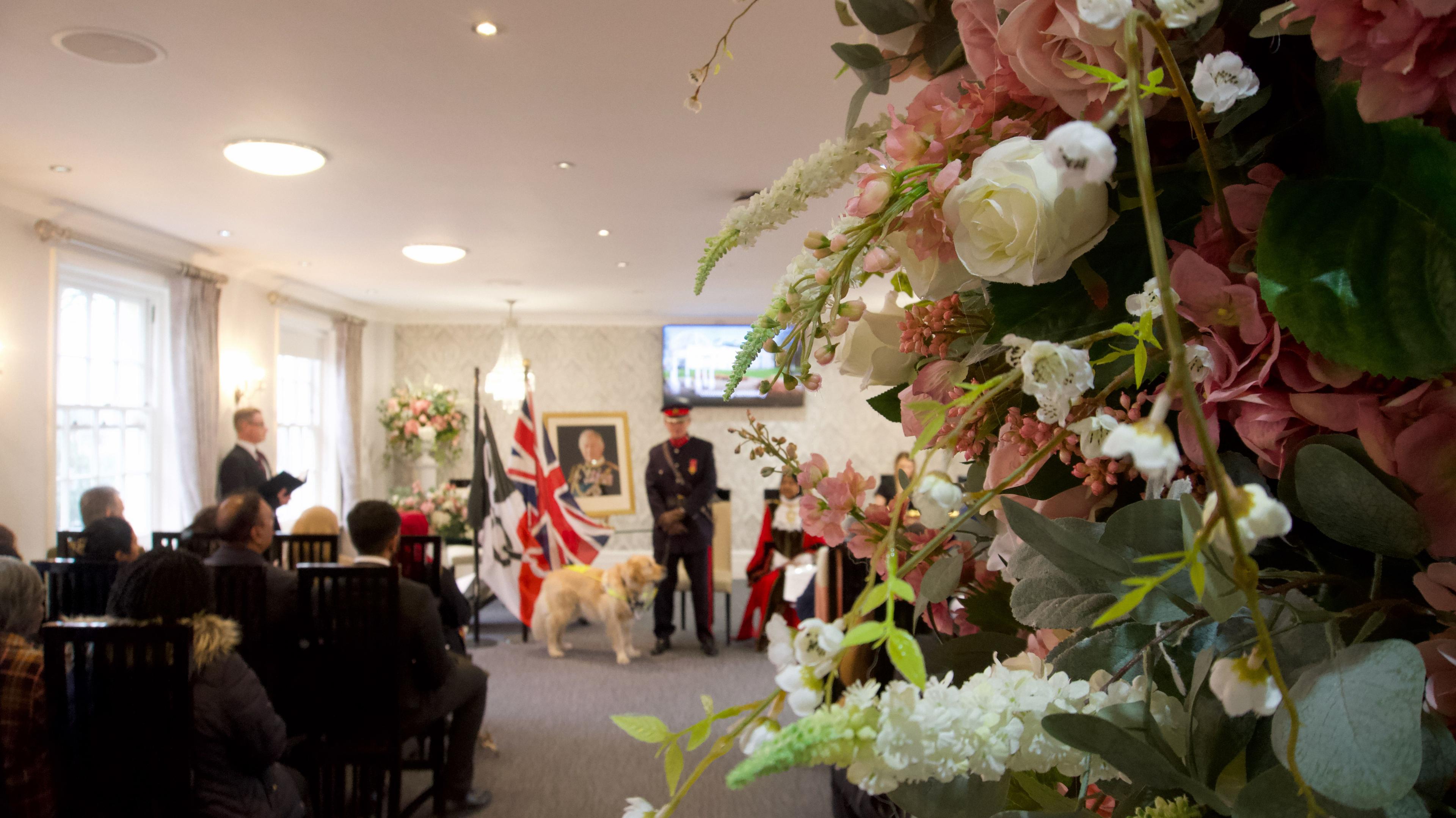 Floral decorations in the foreground - white and pink blooms. In the background, guests at a citizenship ceremony listen to a man in a military uniform with his guide dog