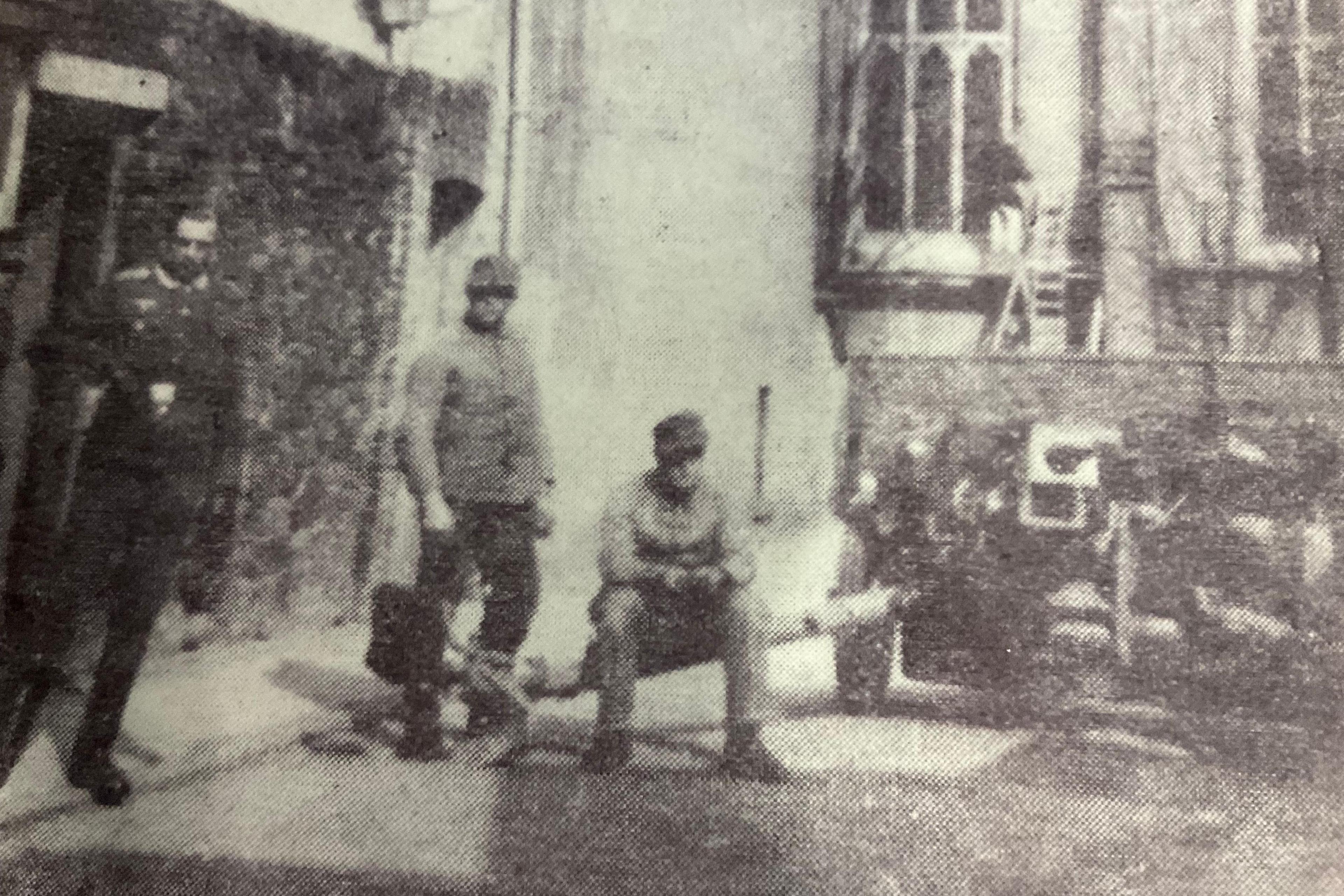 A black and white photo of German soldiers and guns from World War Two on a street corner. The men are all in uniform - one stands next to a doorway in an officer's uniform. Two dressed in ordinary combat uniforms are next to a large anti-tank gun, with one sitting on a part of it.