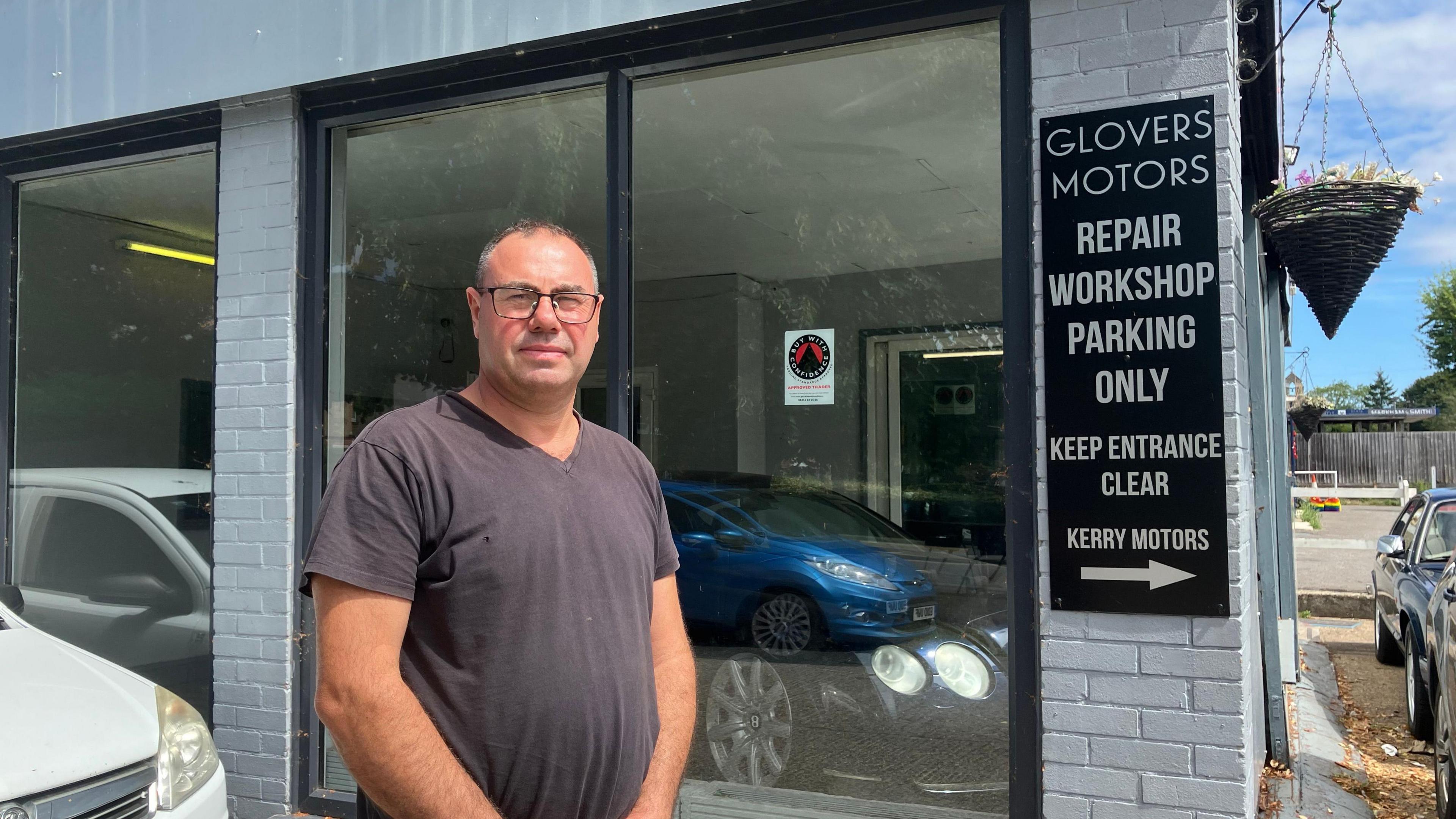 A man with glasses and a grey t shirt standing in front of some shop windows