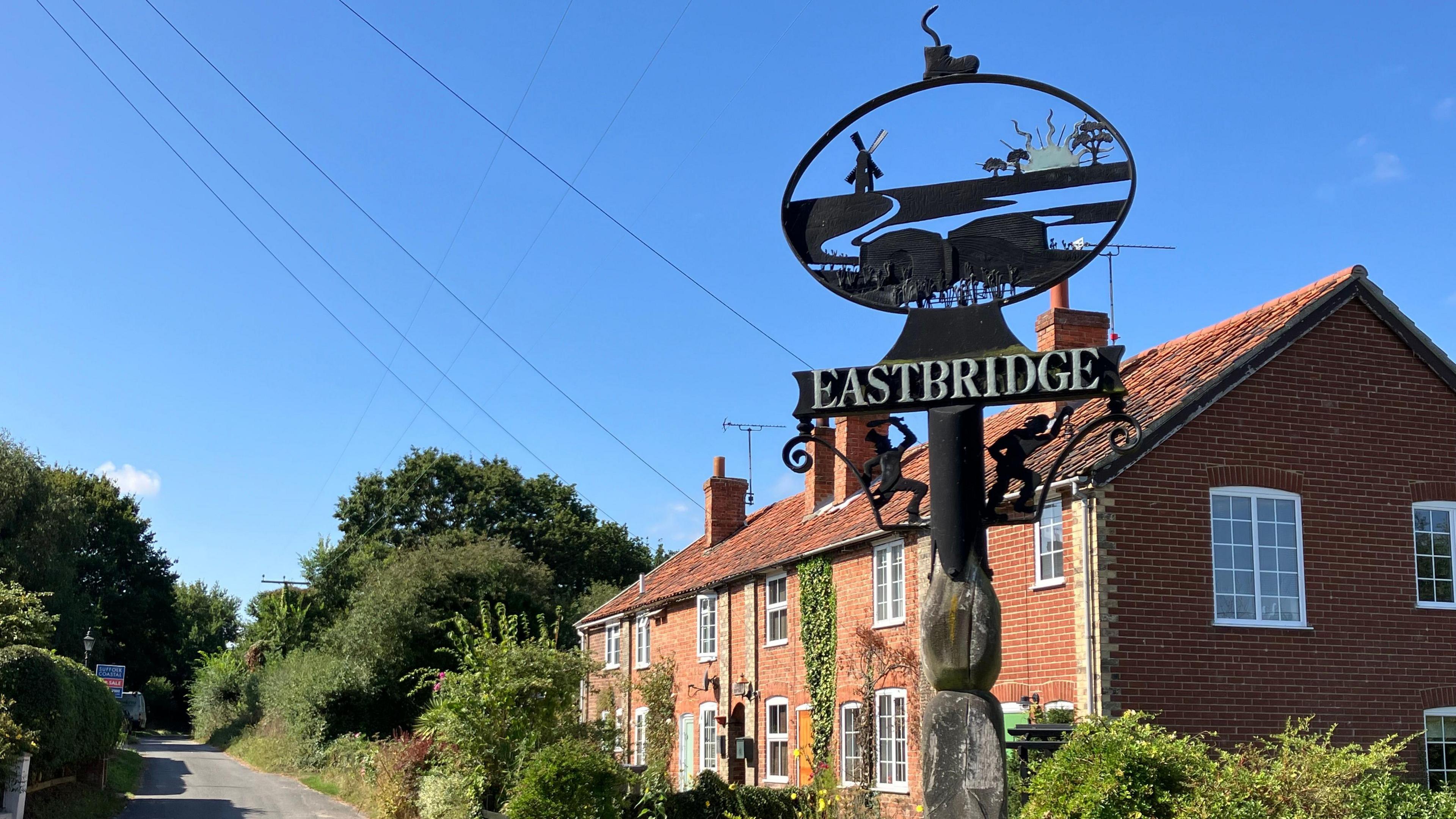 The village sign of Eastbridge depicting a rural scene and a windmill can be seen in front of red-bricked cottages.