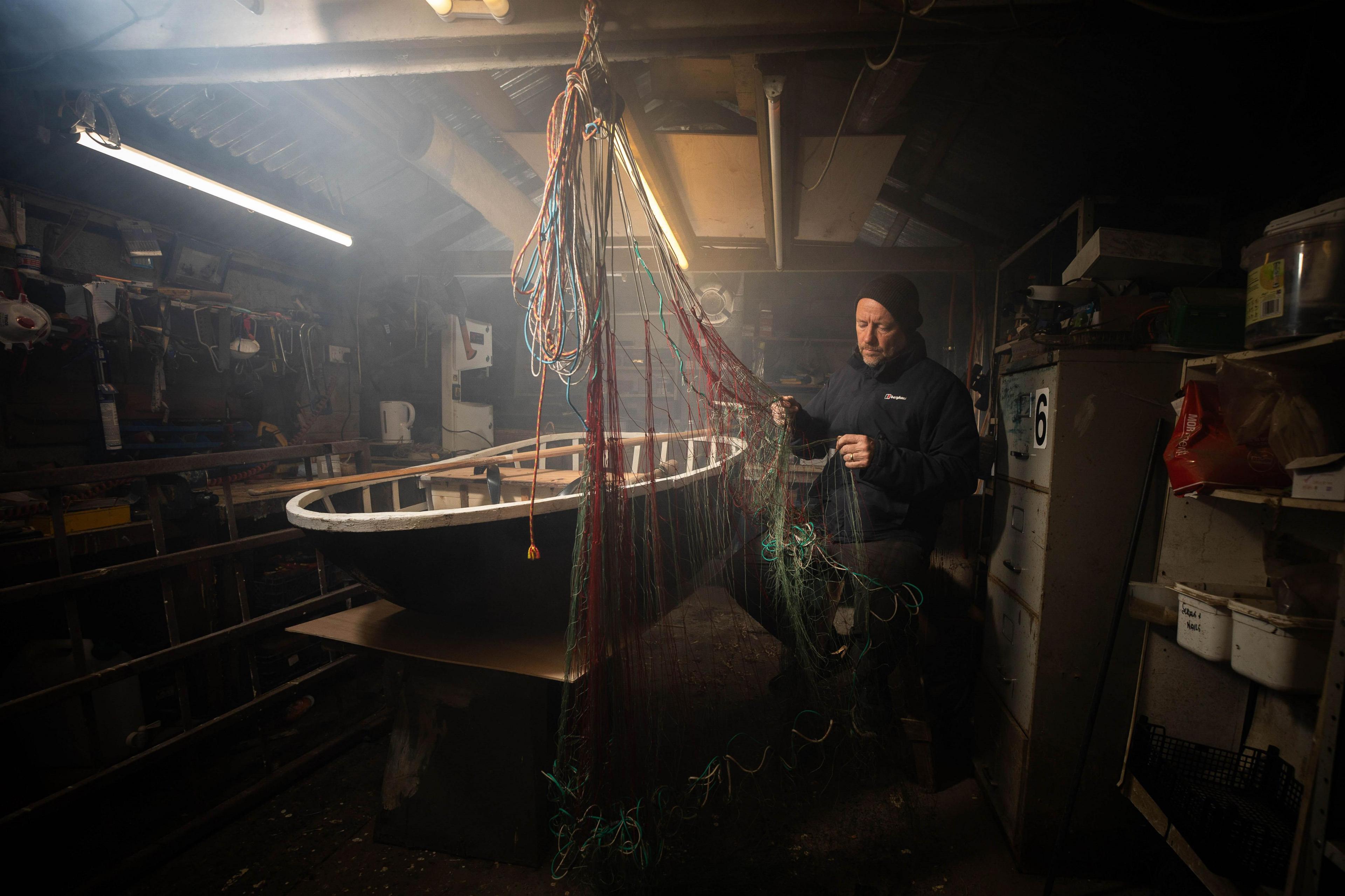 A man in a shed, he is looking at a fishing net and also has it in his hand. A coracle boat can be seen on a table. He has a blue coat and a black beanie hat on.