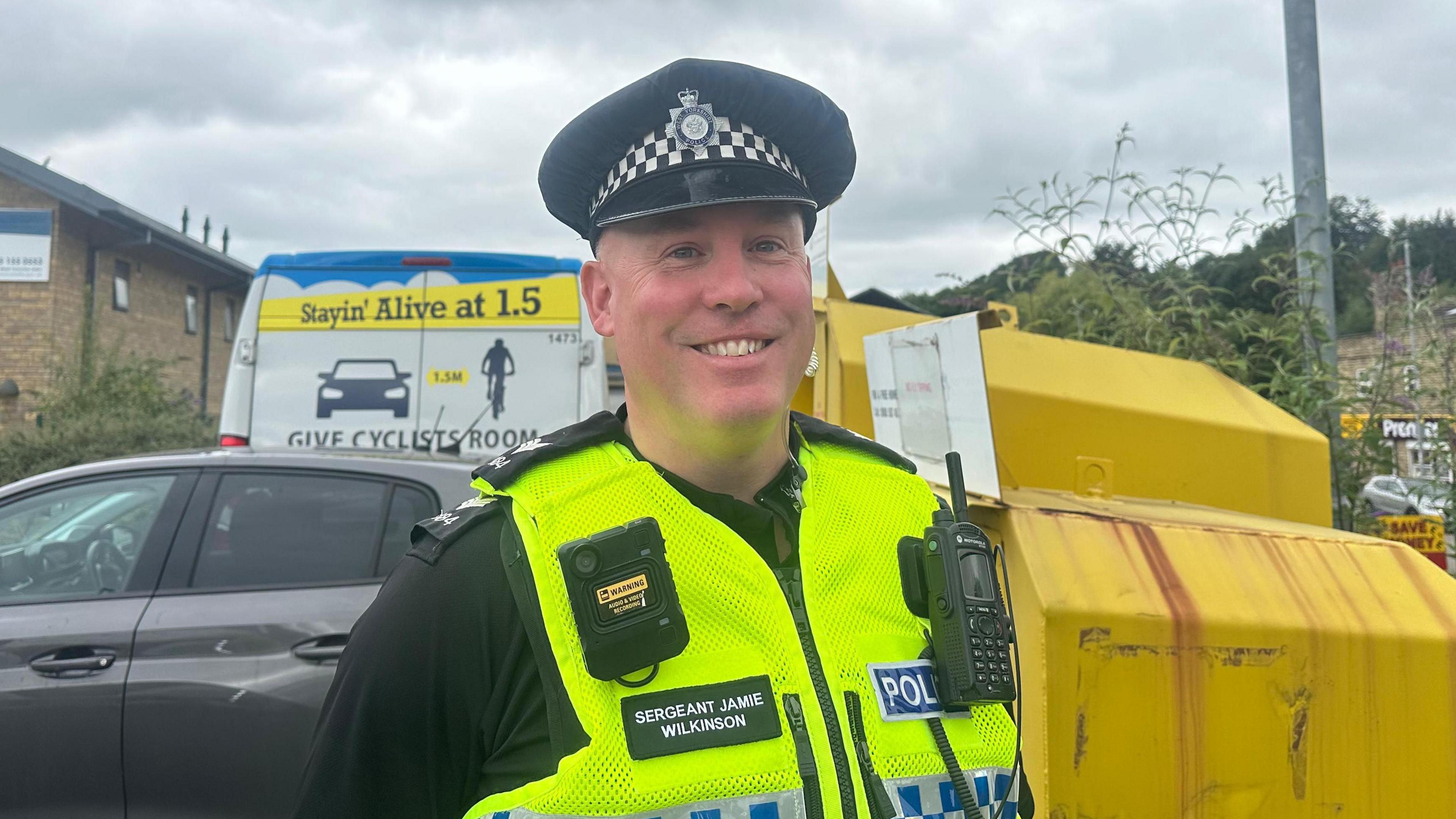 A male police officer in uniform with a bodycam, radio and hat on, smiling. His name badge reads 'Sergeant Jamie Wilkinson'.