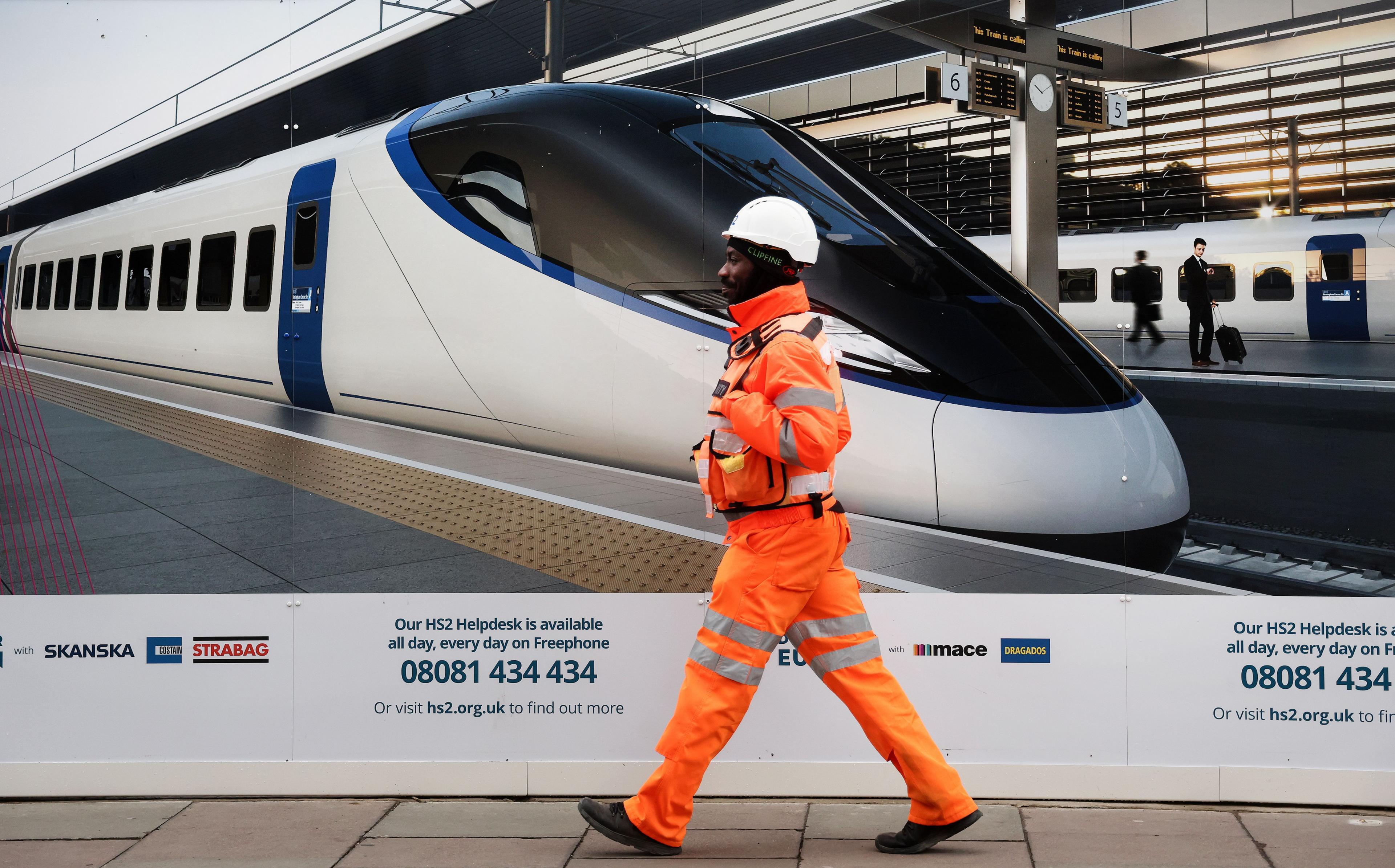 A High Speed 2 worker dressed in orange hi-vis passes a billboard of a HS2 train at the Euston terminal.