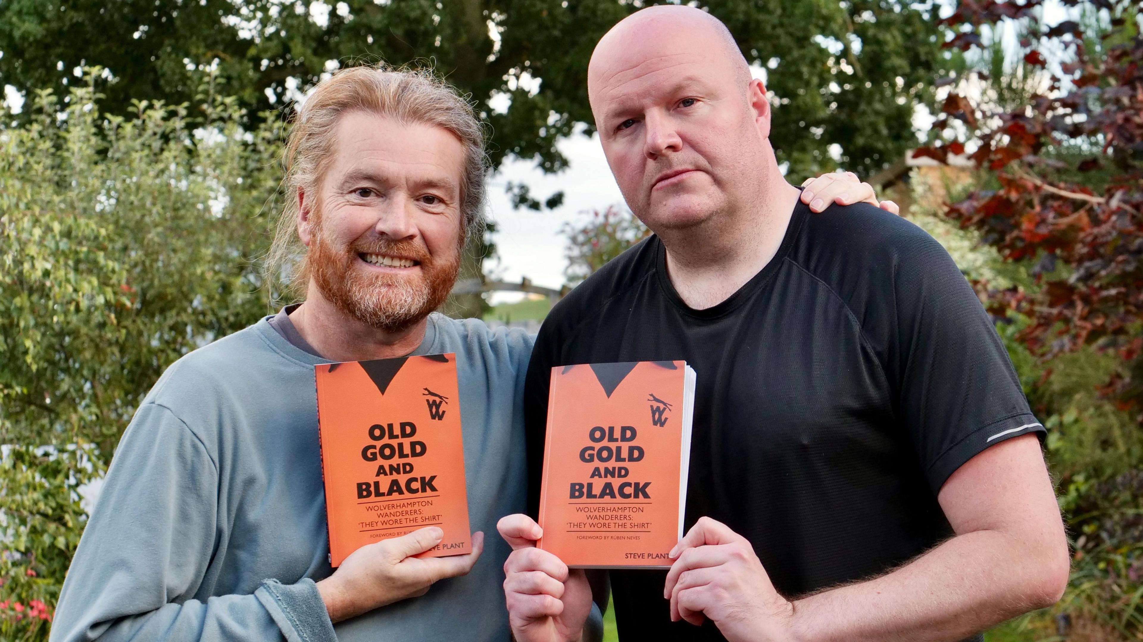 Two men in a garden both holding copies of a book called Old Gold and Black 