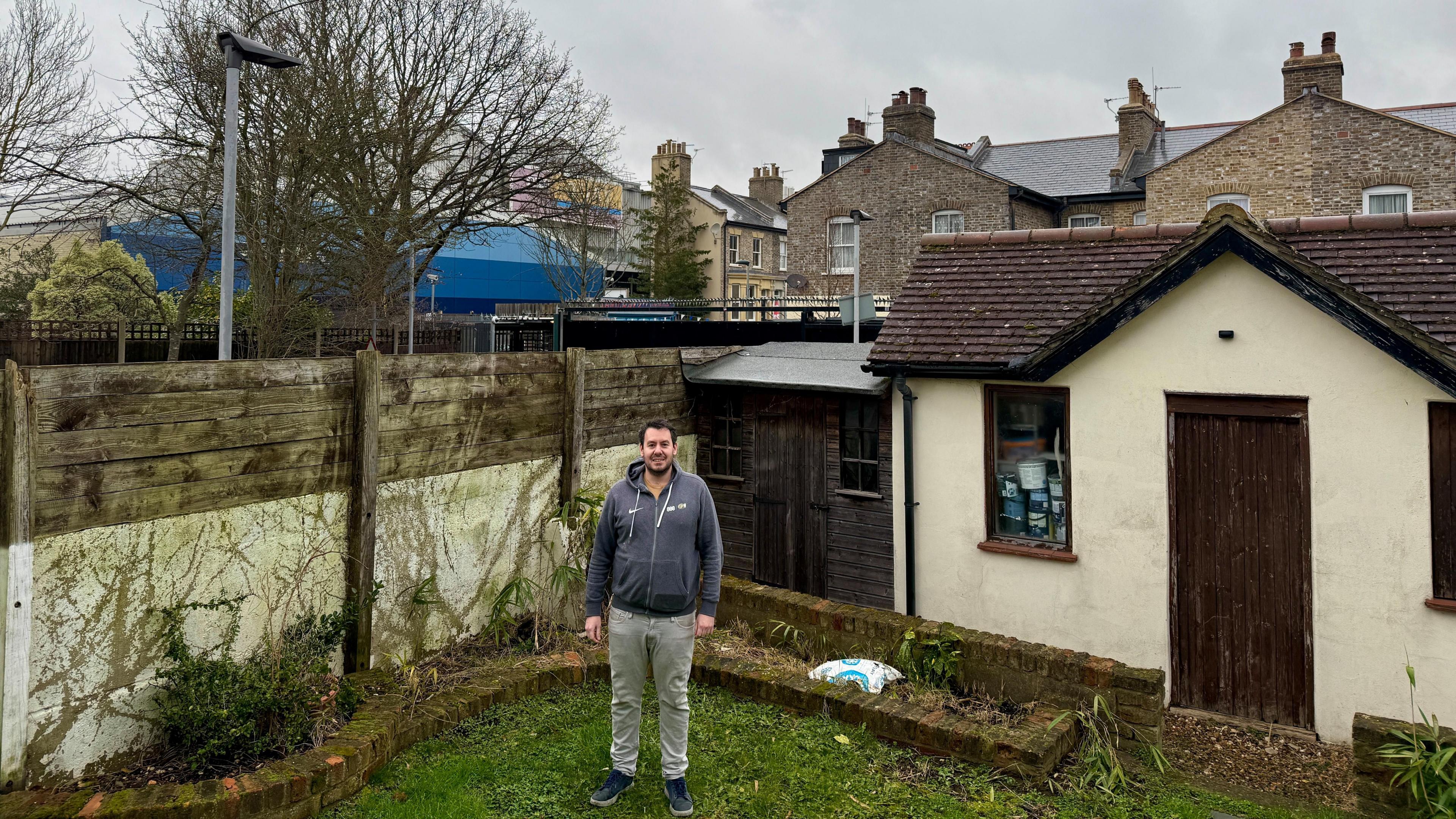 A man with short brown hair and a beard is stood in his back garden. Behind him you can see see part of the EastEnders set.