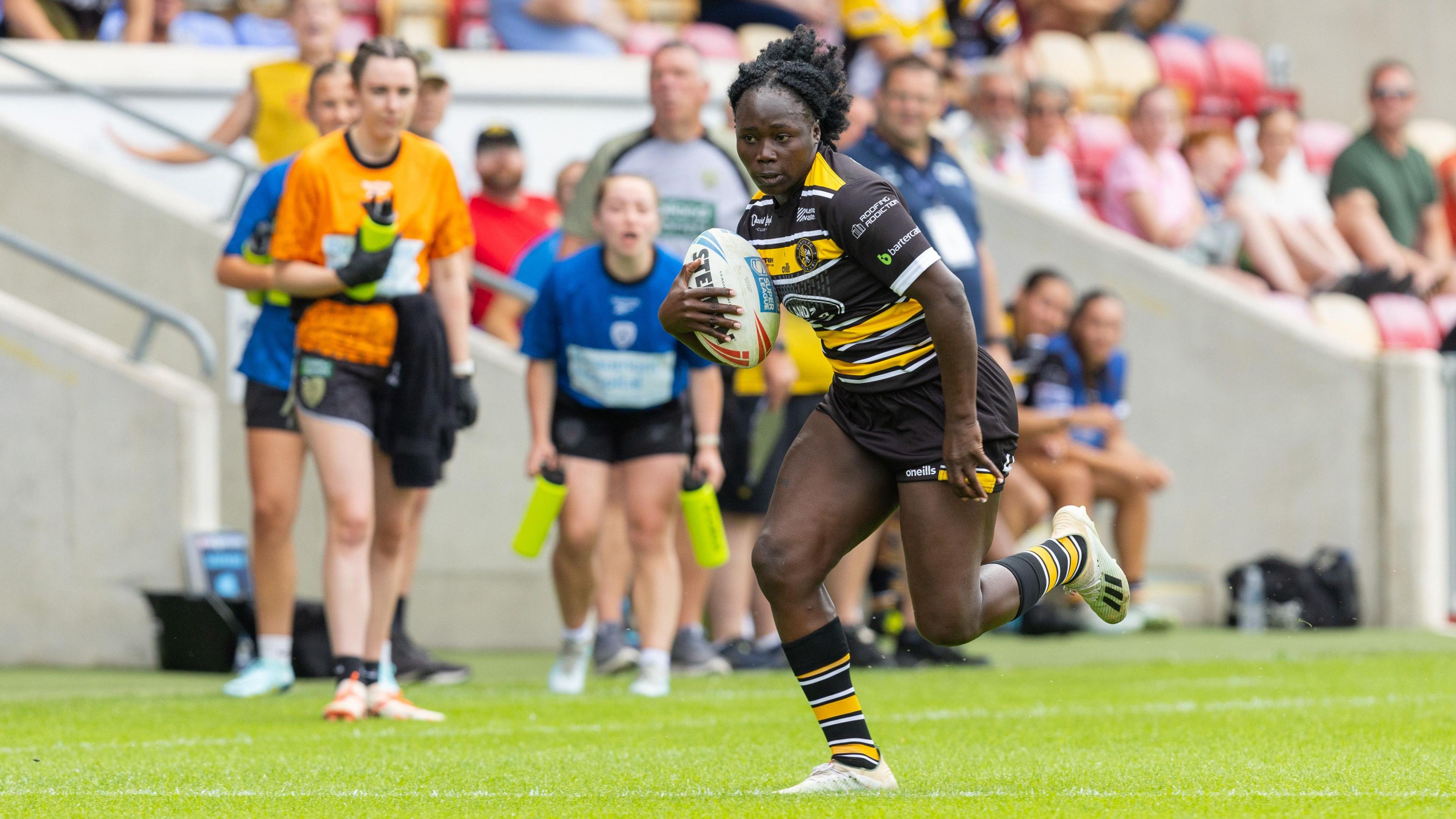 Peace Lekuru in action on the rugby pitch, running with the rugby ball. She wears a York Valkyrie rugby shirt which is black with a yellow stripe across the chest, black shorts, and black and yellow striped socks. Spectators can be seen behind her in the stands, and teammates watch on.