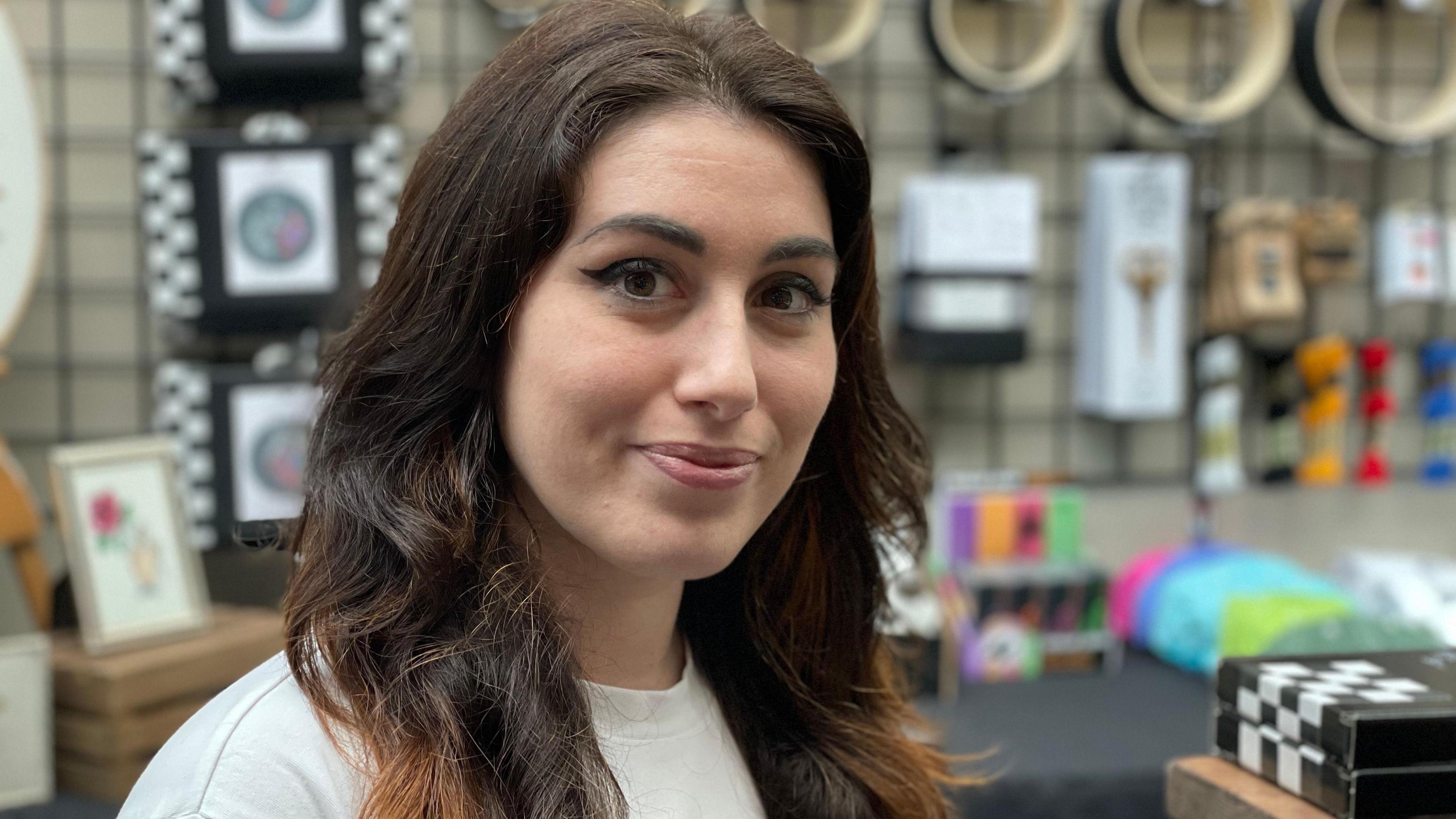 A young woman smiles at the camera. She has dark hair and dark eyes. Her stall is behind her and a range of products are visible on the shelves.