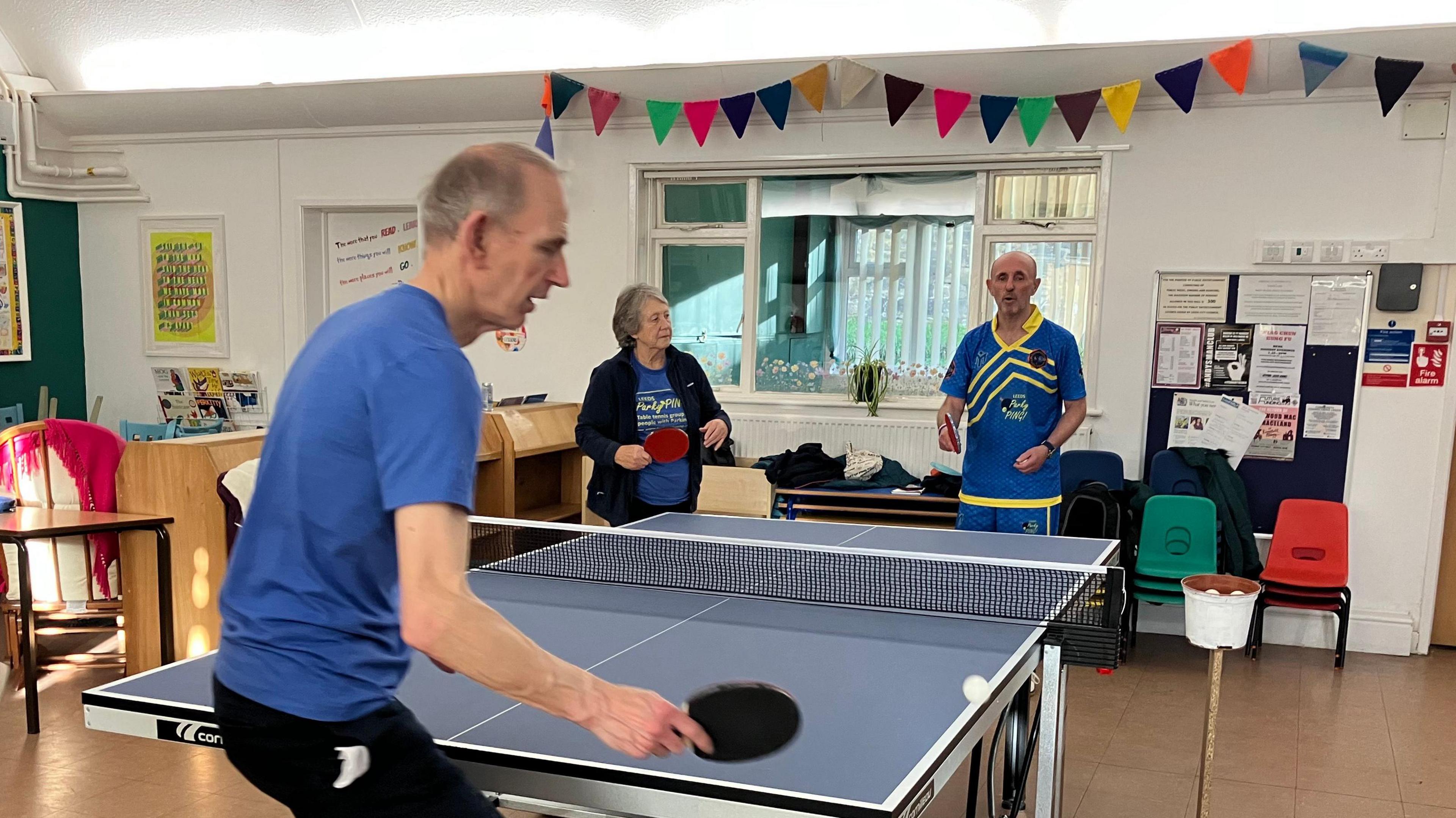 Three people are gathered around an indoor table tennis setup. One person in the foreground, wearing a blue shirt, is facing the table. Two others stand on the opposite side, one holding a red paddle wearing a black fleece and the other wearing a blue and yellow sports jersey.
