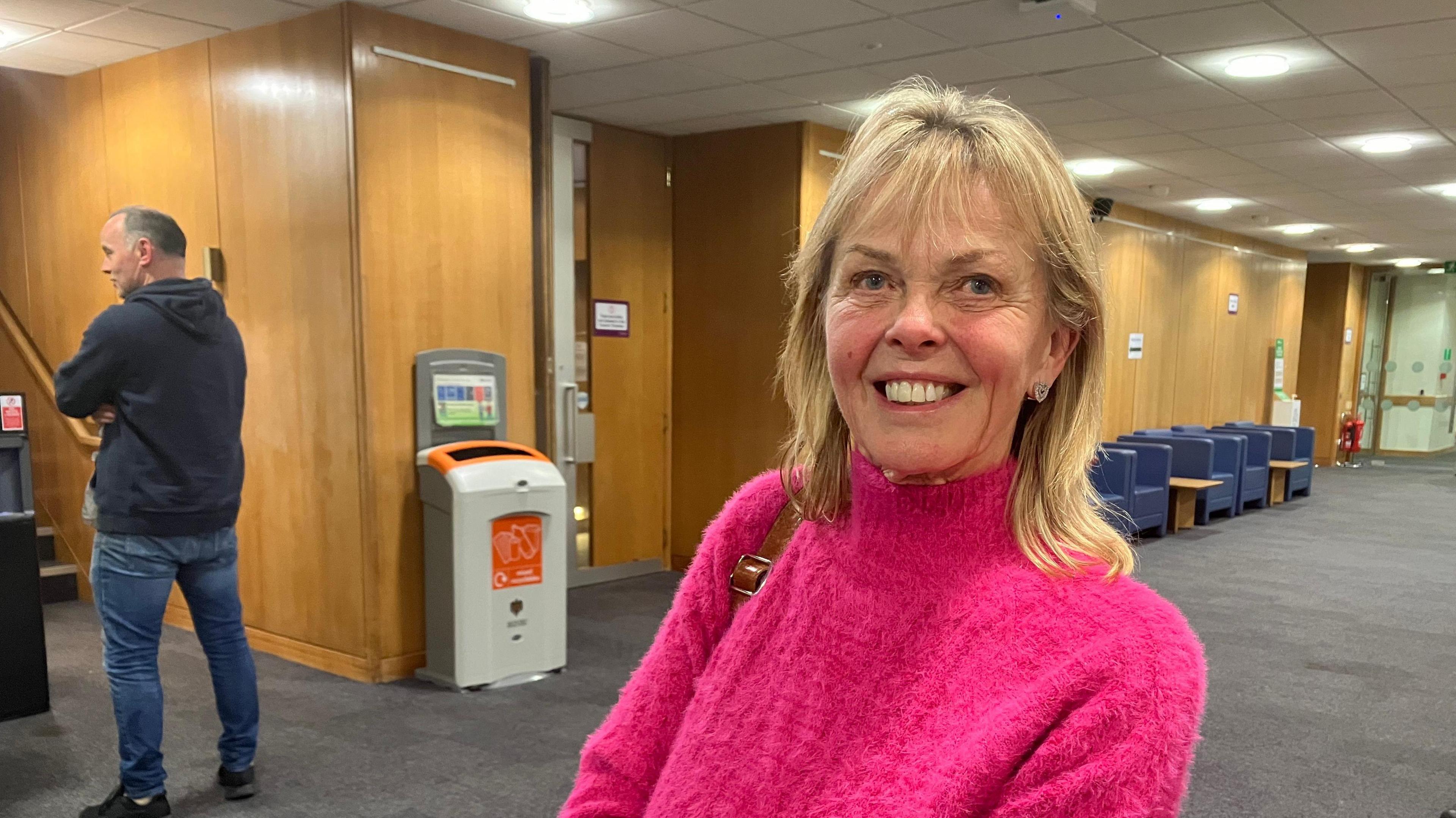 A picture of a blonde haired woman with shoulder length hair, she is wearing a pink roll neck jumper.