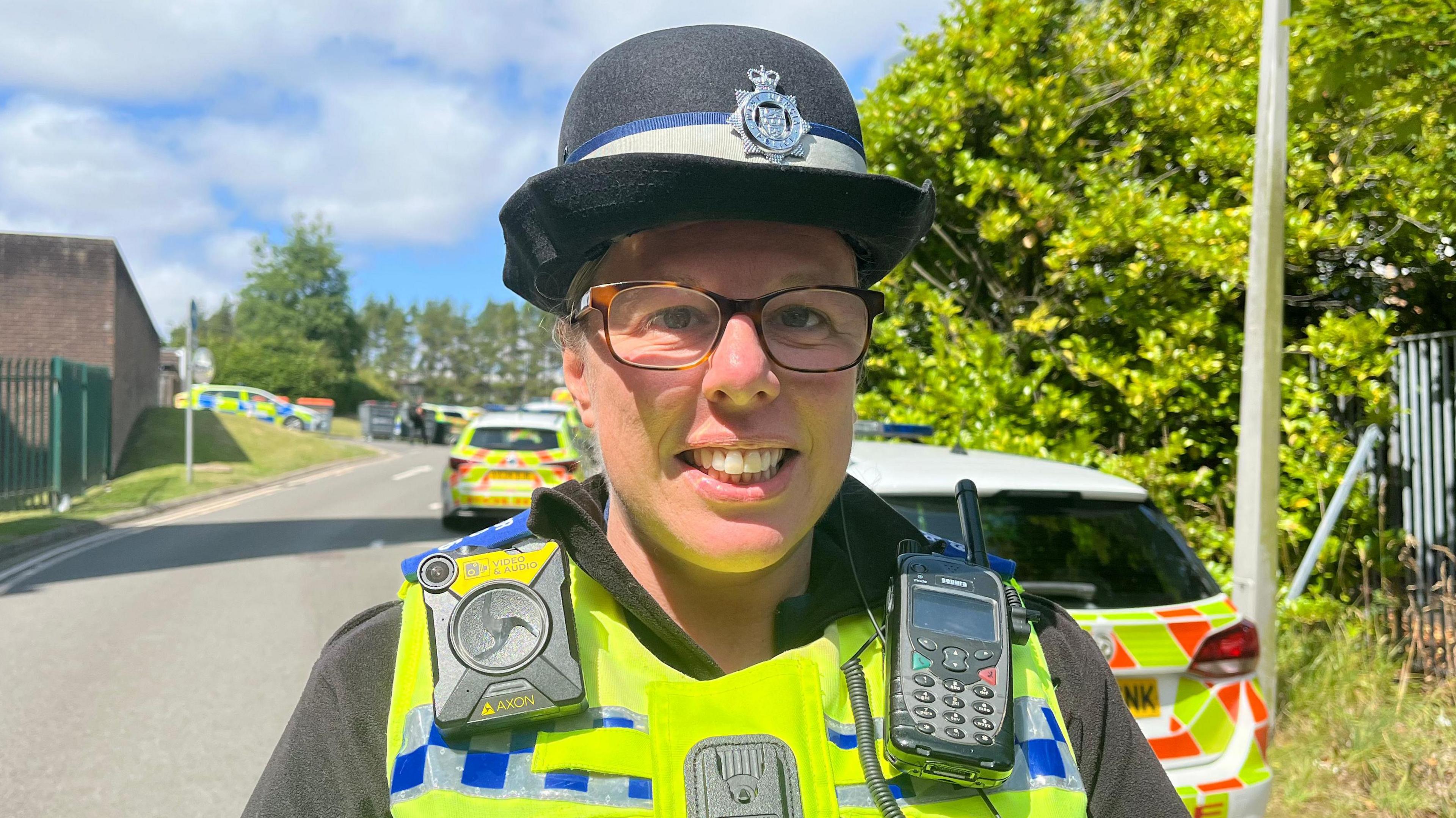 Katy is looking into the camera and smiling. She's wearing a black fleece with a bright yellow reflective vest over the top. On her left shoulder is a police radio, and on her right shoulder is a body worn camera. She's also wearing a police hat. The sky is blue and there's lots of sunshine, and she's stood in front of a police car. 