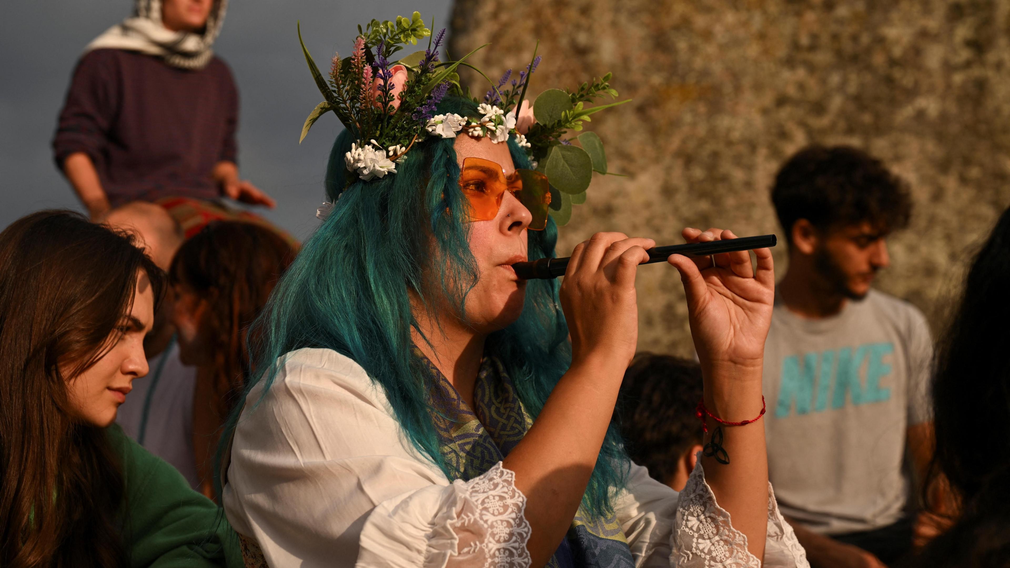 A person wearing a flower crown with blue hair and orange sunglasses. They're playing a small flute with Stonehenge and people behind them