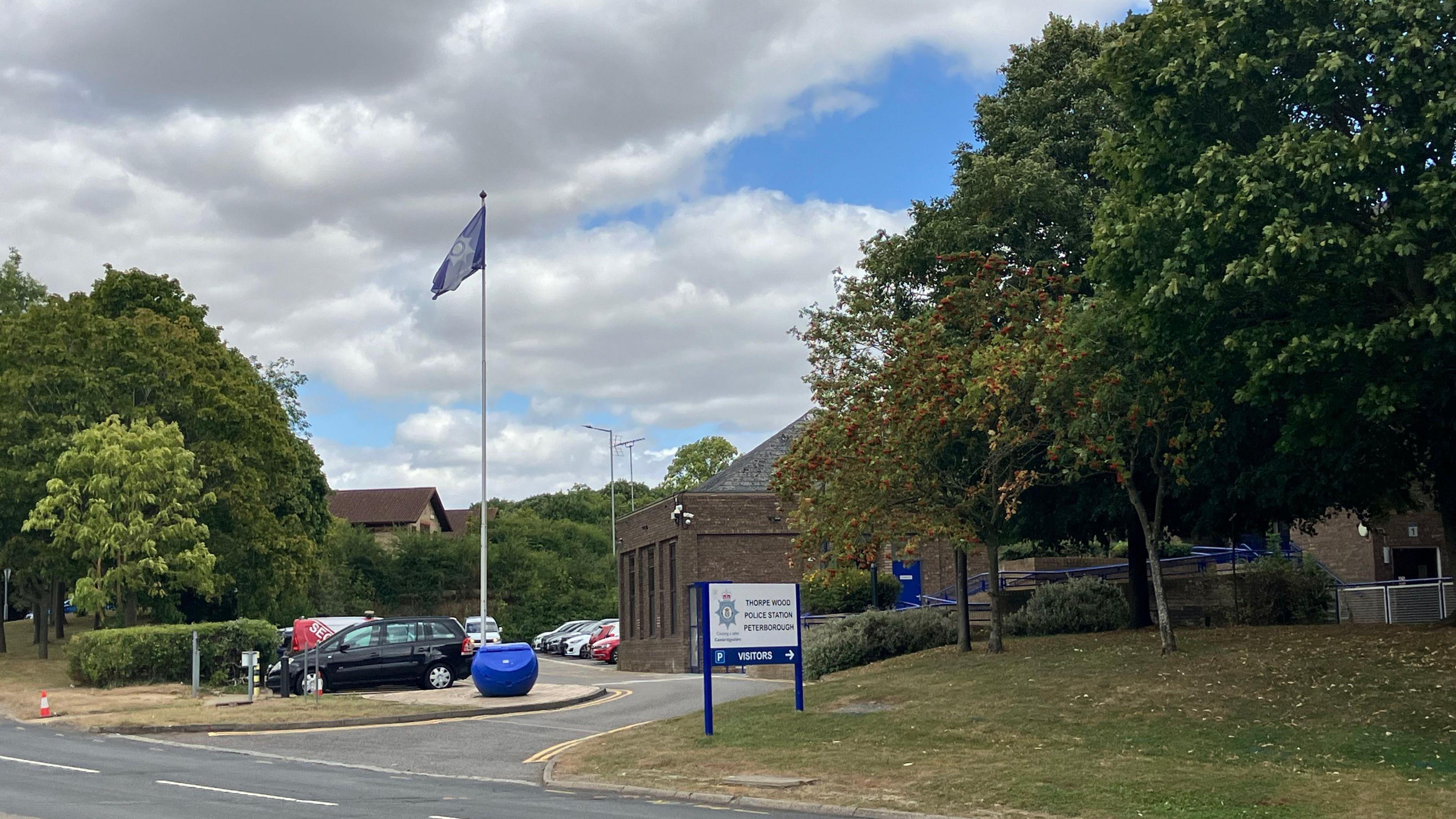 Street view of Thorpe Wood Police Station. There is a flag pole with a flag with the police emblem on it at the top and a white sign  with Thorpe Wood Police Station written on it.