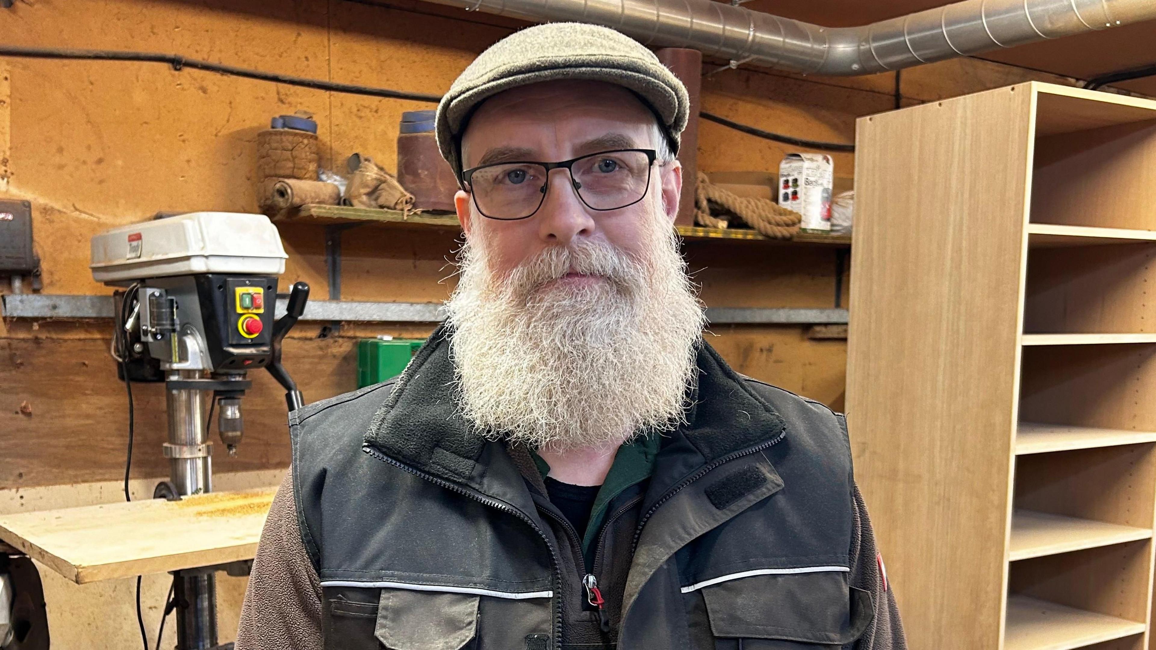 Mark Lucas is seen in a flat cap and glasses stands in a woodworking shop, surrounded by tools and wooden shelving units.
