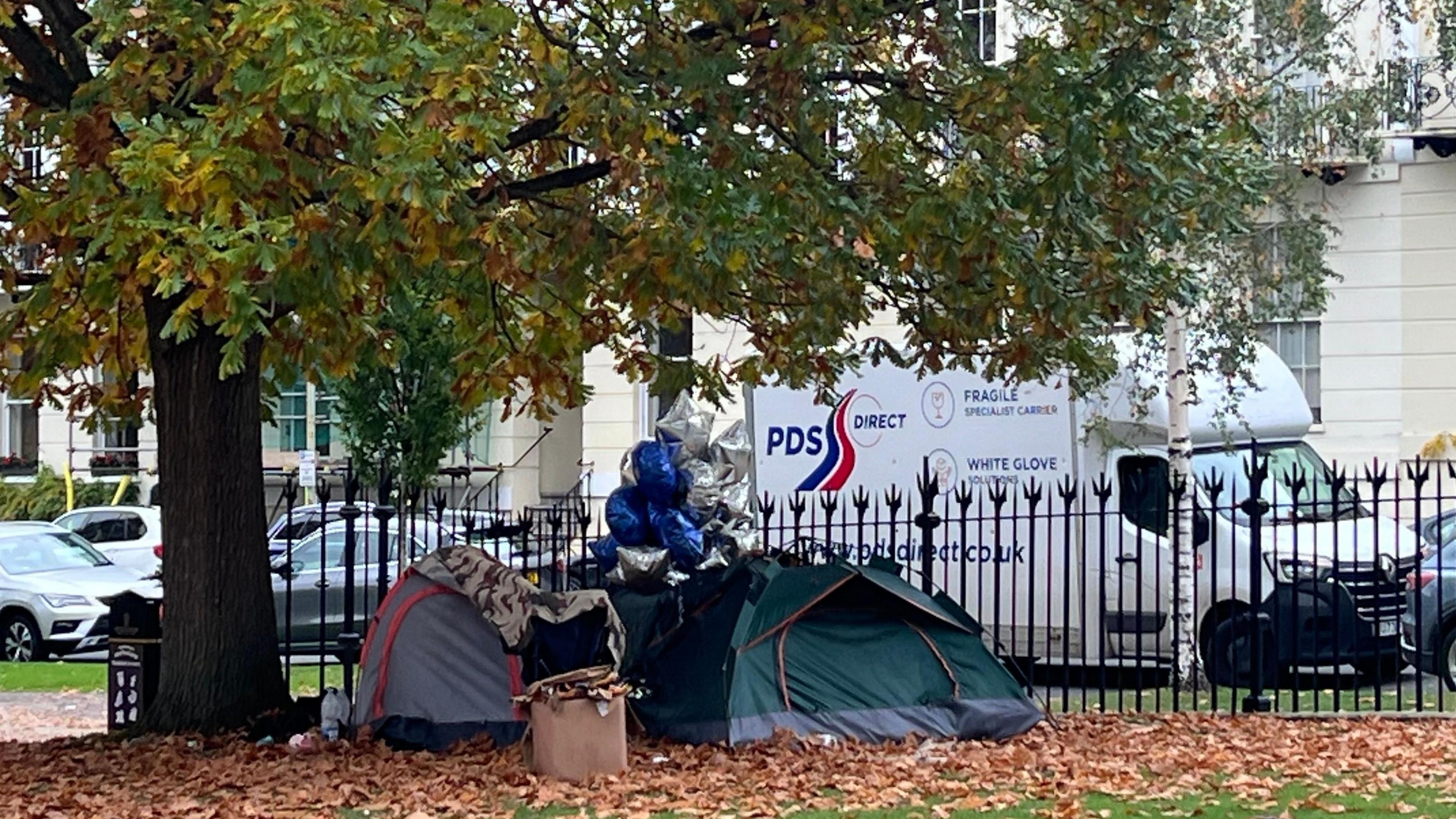 Three tents, surrounded by leaves, a bunch of blue and silver balloons, and a washing basket, sit beneath a tree at the edge of a Cheltenham park, bordered by wrought iron fencing. White Regency style terraced homes can be seen on the other side of the road, where cars and a van are parked.