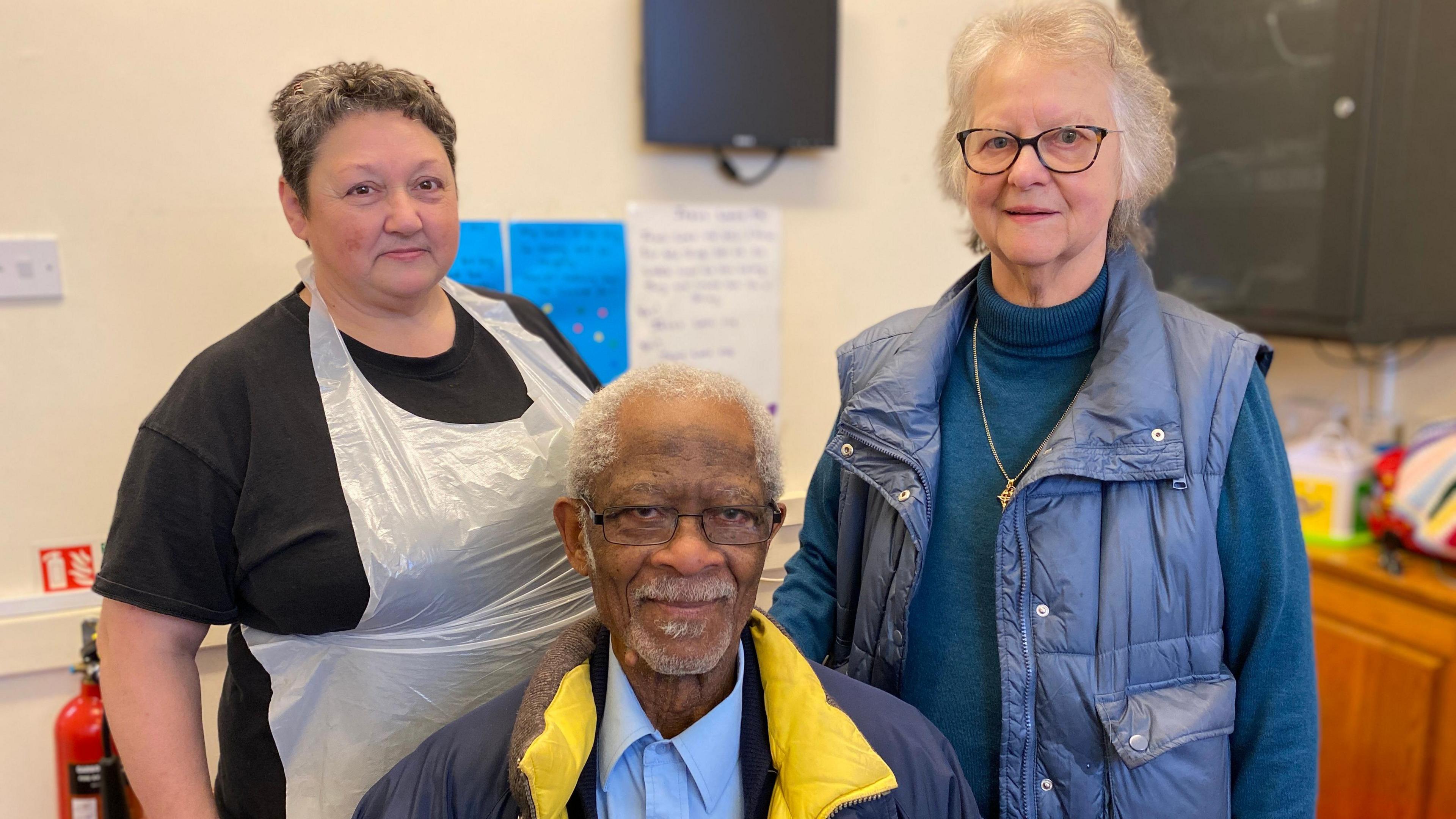 Claudia, Clement and Donna are standing next to each other and smiling. Claudia has short brown hair, is wearing a black t-shirt and an apron. Clement is wearing a yellow and blue puffer coat and glasses. Donna has a blue jumper and gilet, and has black glasses.