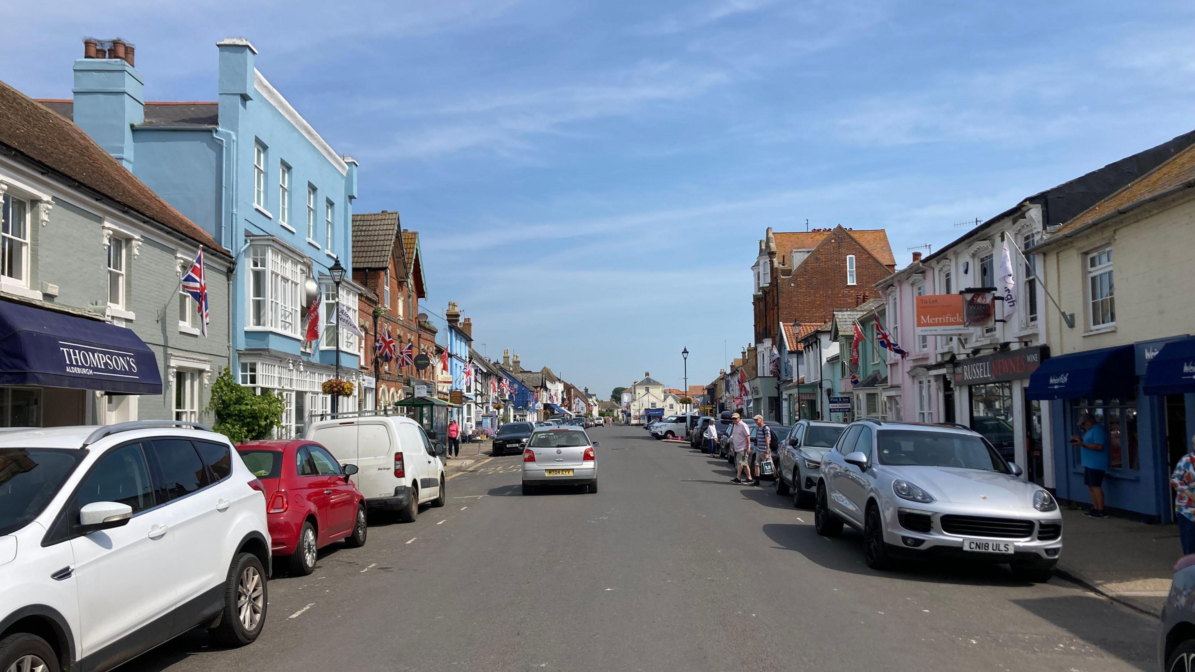 A view of a road in Aldeburgh. Colourful buildings line each side of the road and cars are parked on either side.