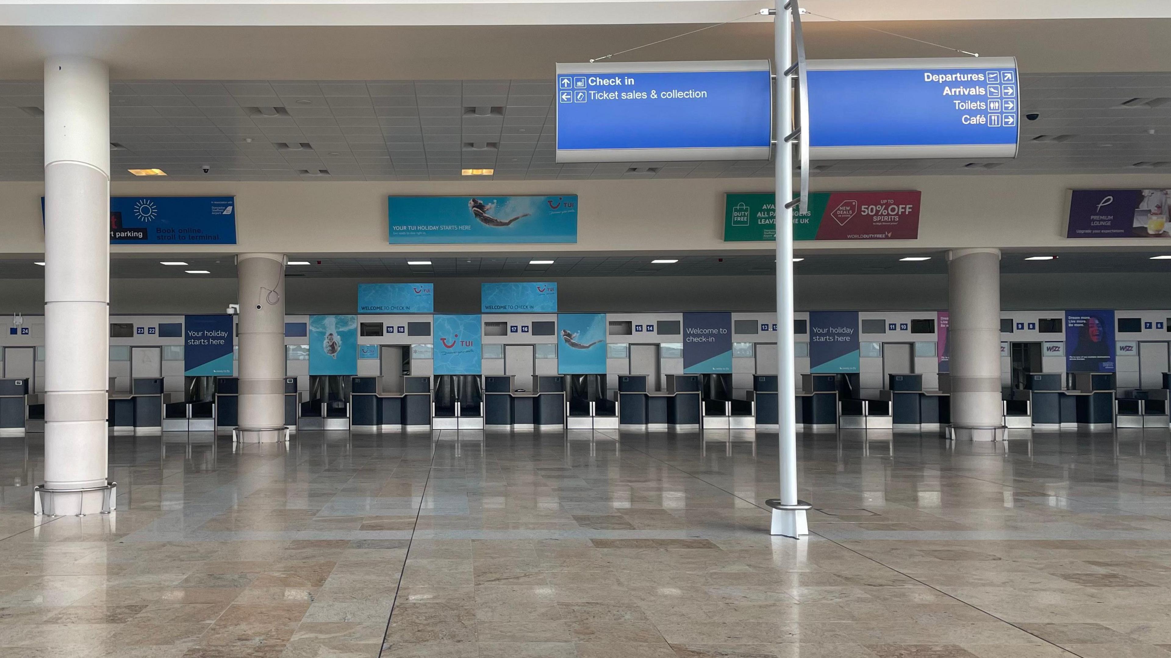 An empty airport check-in area is shown, with rows of TUI-branded desks lining the back wall. In the foreground, a signpost directs passengers to ticket sales, departures, arrivals, toilets, and a cafe.