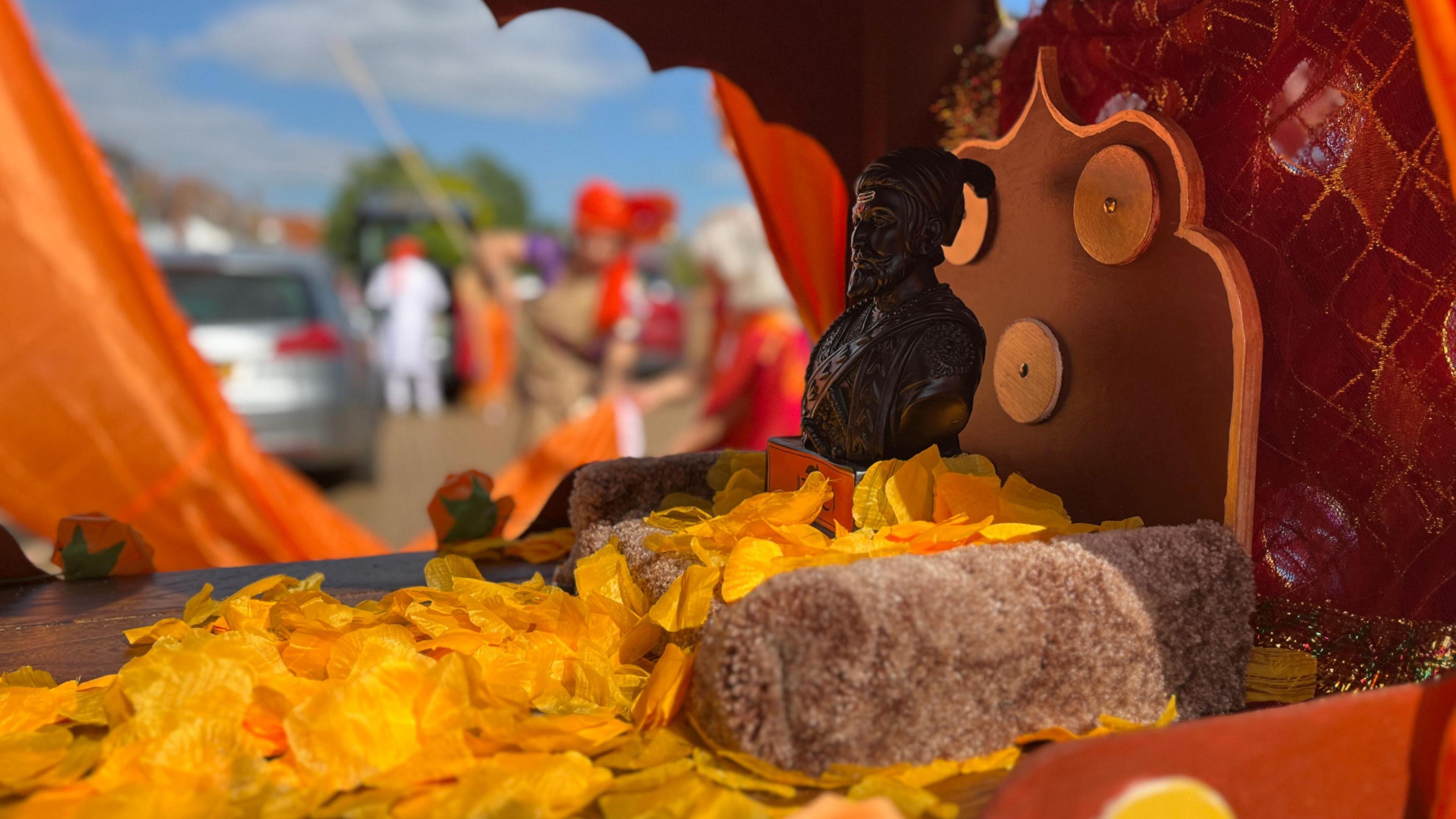 A small bronze statue of a Hindu king sits on a wooden carrier with orange petals