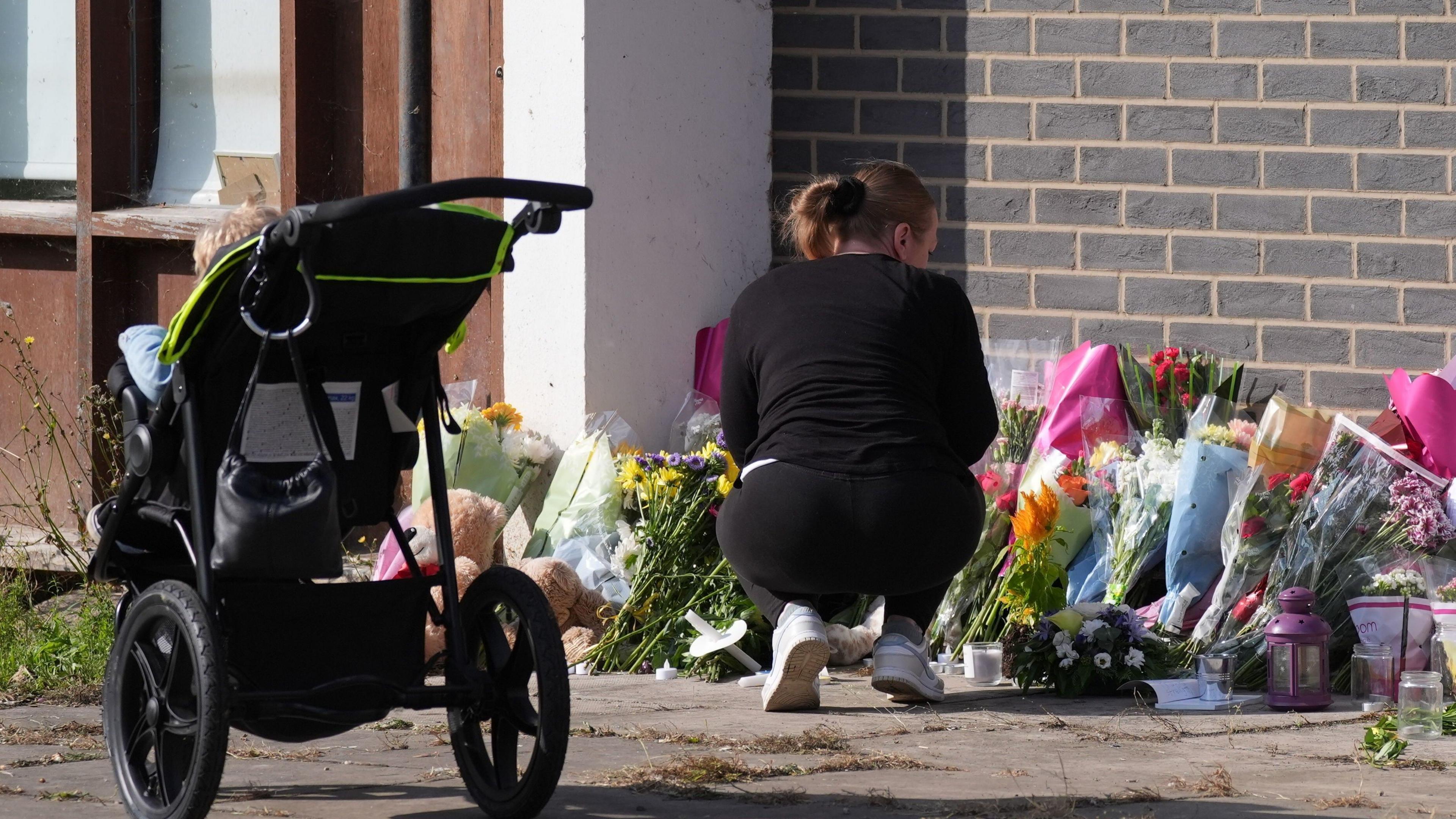 A woman crouches down in front of a number of bunches of flowers propped against a breeze-block wall. She is wearing black sportswear and white trained. A pushchair stands nearby.