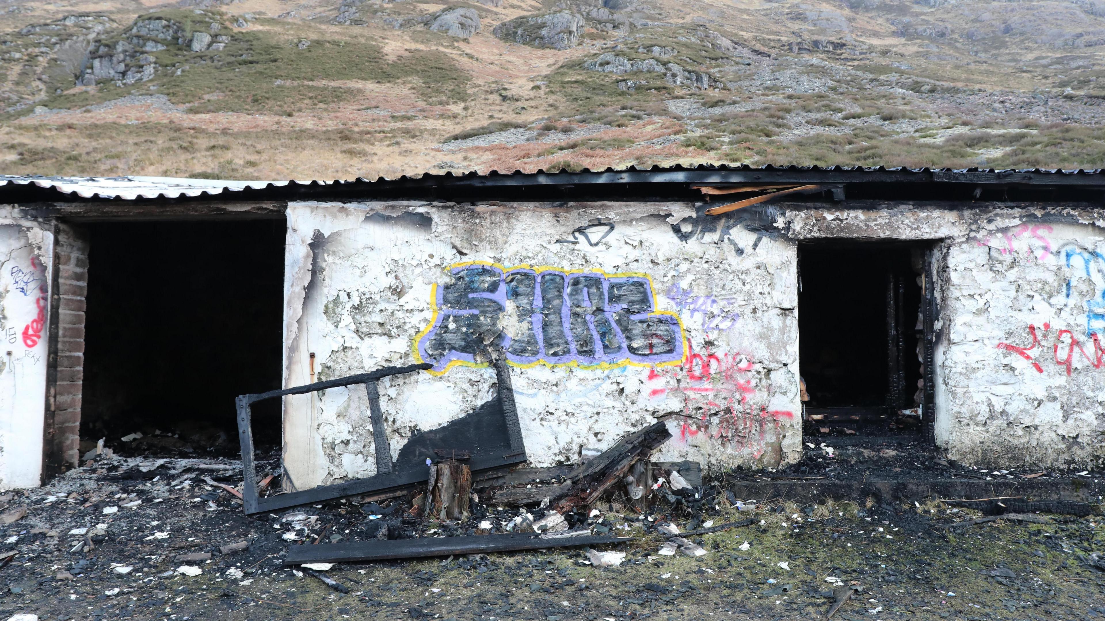 Exterior of an outbuilding at the former Savile property at Allt Na Reigh - a white stone building with black tin roof, heavily covered in graffiti with fire damage on the exterior and slightly visible on the interior. There are fire damaged pieces of wood - possibly window frames - leaning against the wall with debris on the ground