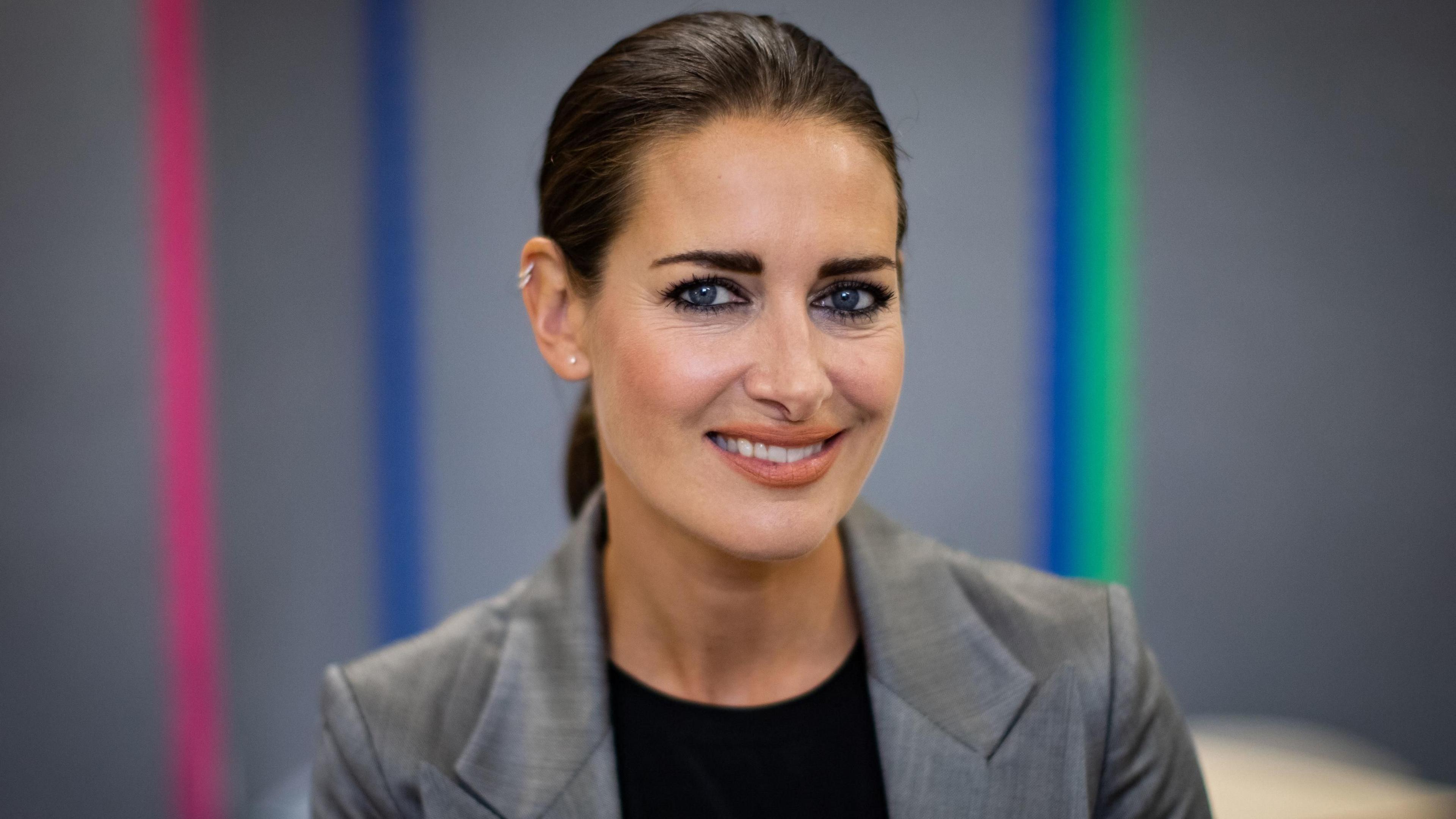 Kirsty is wearing a grey blazer and a black top, smiling directly at the camera. Her hair is tucked neatly in a ponytail and is wearing dark eye shadow which is complimenting her blue eyes and a nude lipstick.