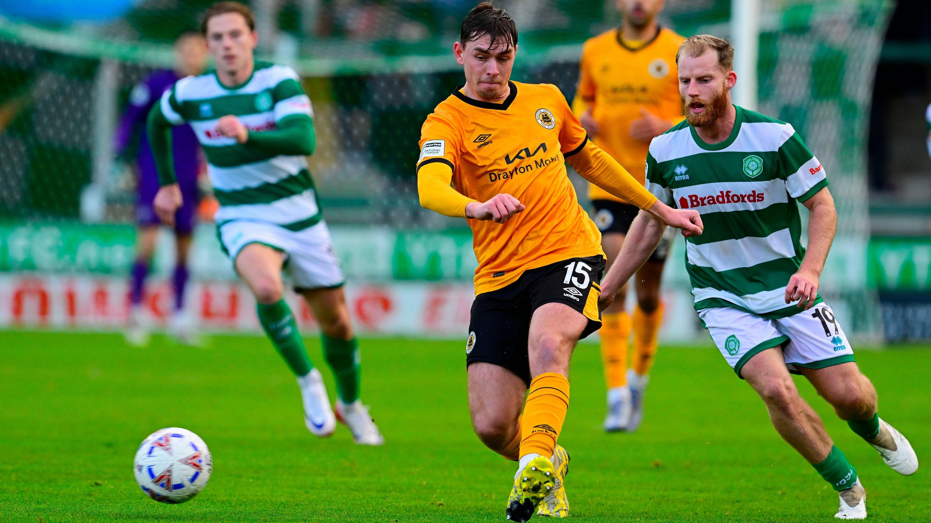 Matty Carson in his orange Boston United kit passes the ball under pressure from a Yeovil Town player wearing a green and white hooped shirt. There is another Yeovil player in the background and another Boston one in the distance