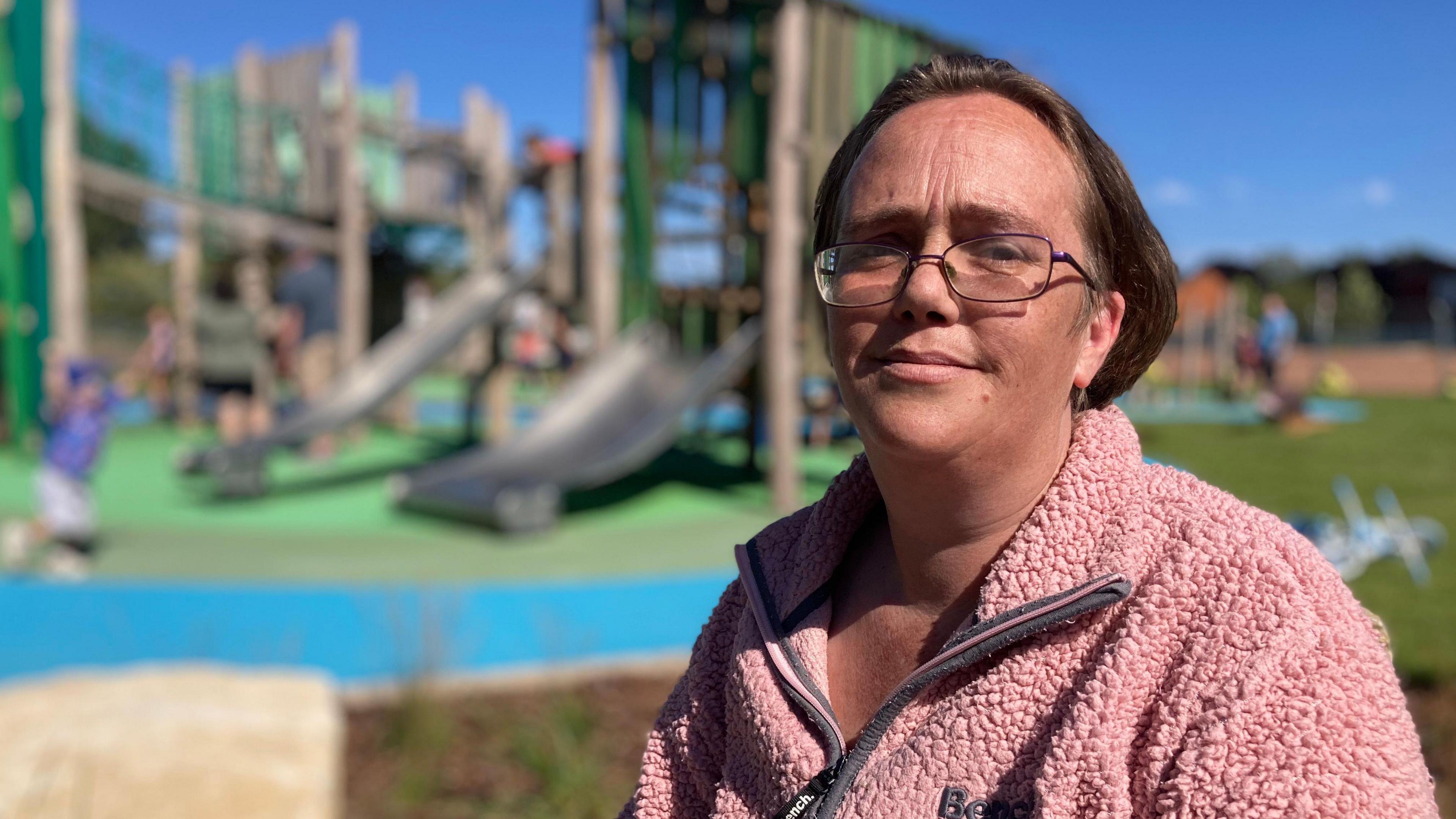 In the foreground, a woman wearing glasses and a pink zipper jacket looks at the camera with a children's play park obscured in the background.