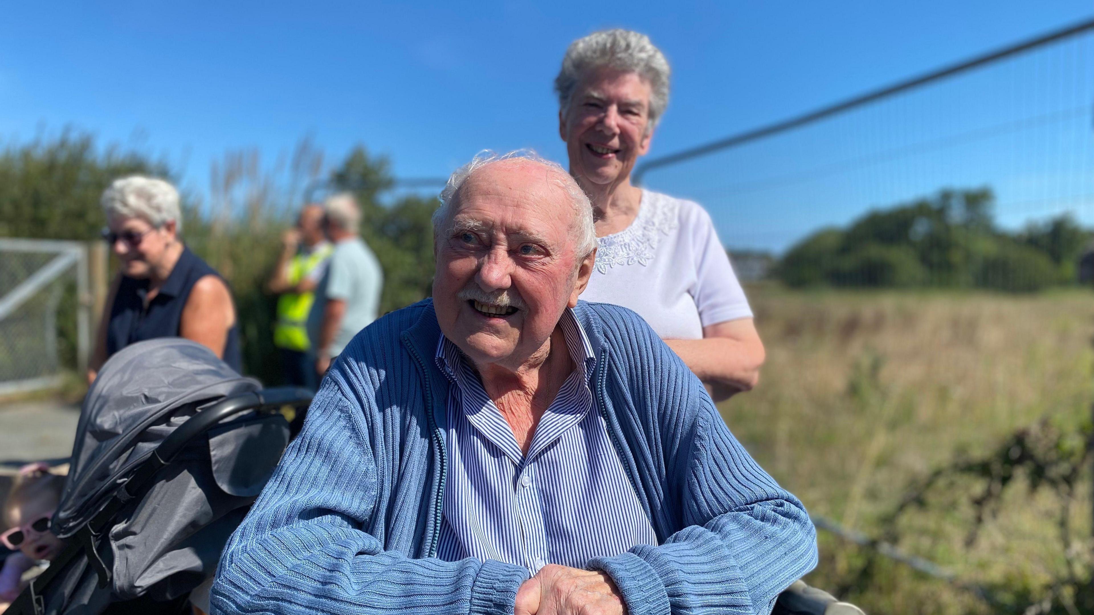 An elderly man sitting in a wheelchair wearing a blue shirt and cardigan. A woman standing behind him is wearing a white blouse.