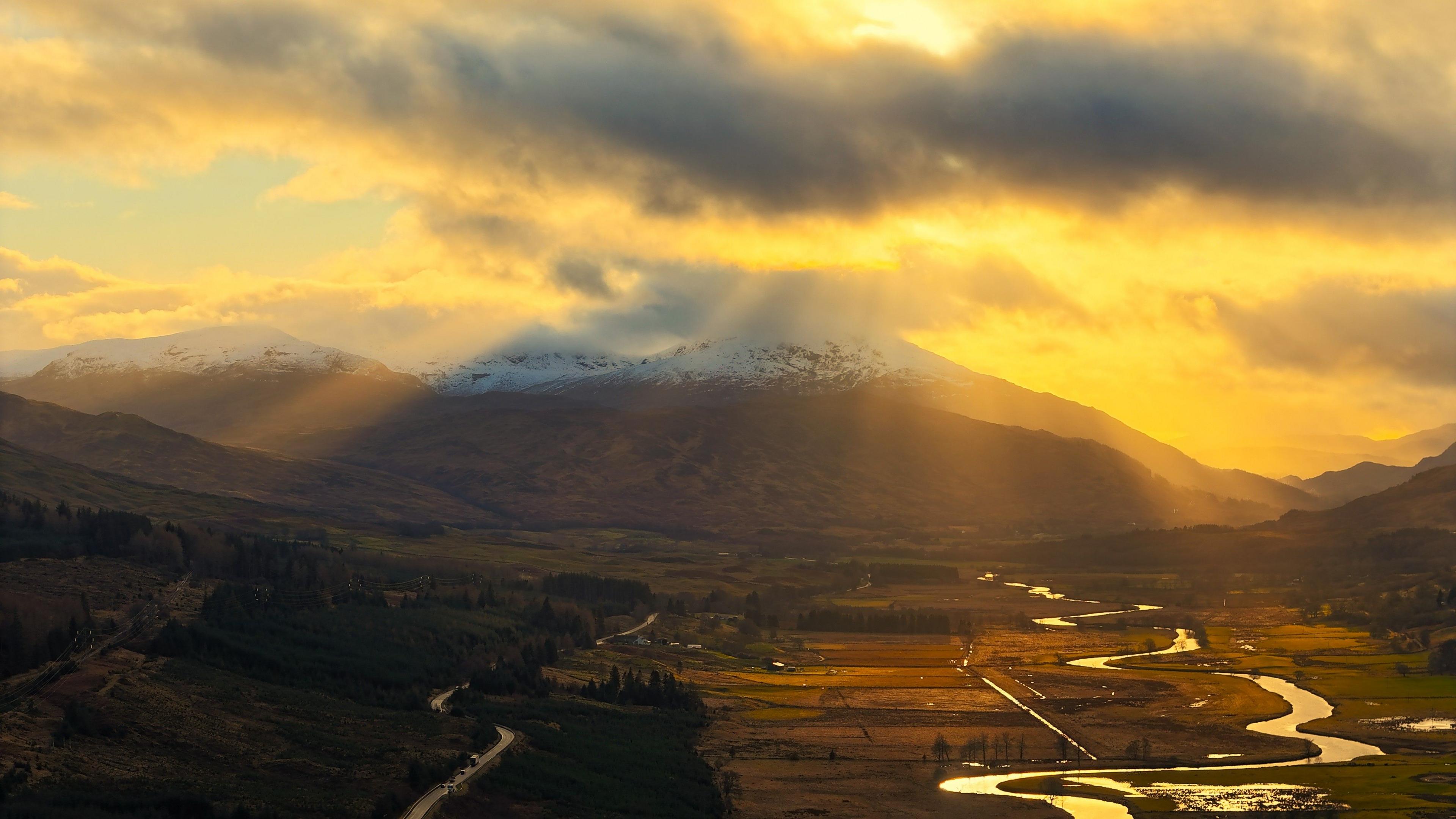 Landscape image of the sun peeking through a cloudy sky, setting an orange glow over mountains and fields with a river snaking through them