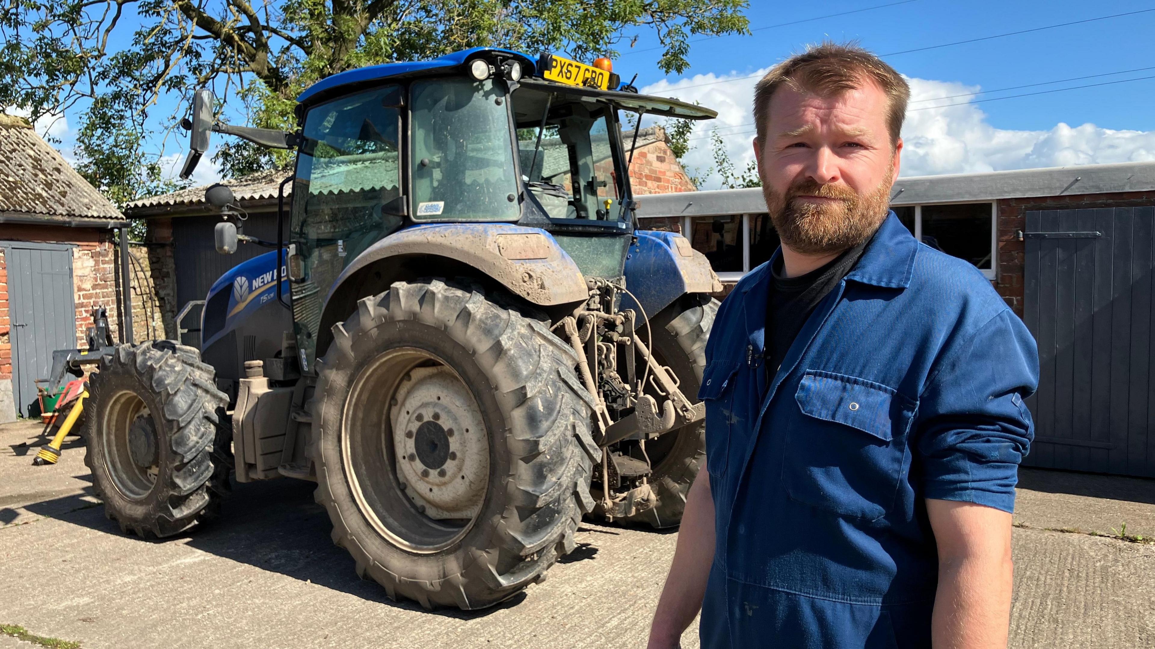 John Story is wearing blue overalls and standing in front of a blue tractor in a farmyard. He has short hair and a beard and moustache. Behind him are low level farm buildings with a tree behind them.