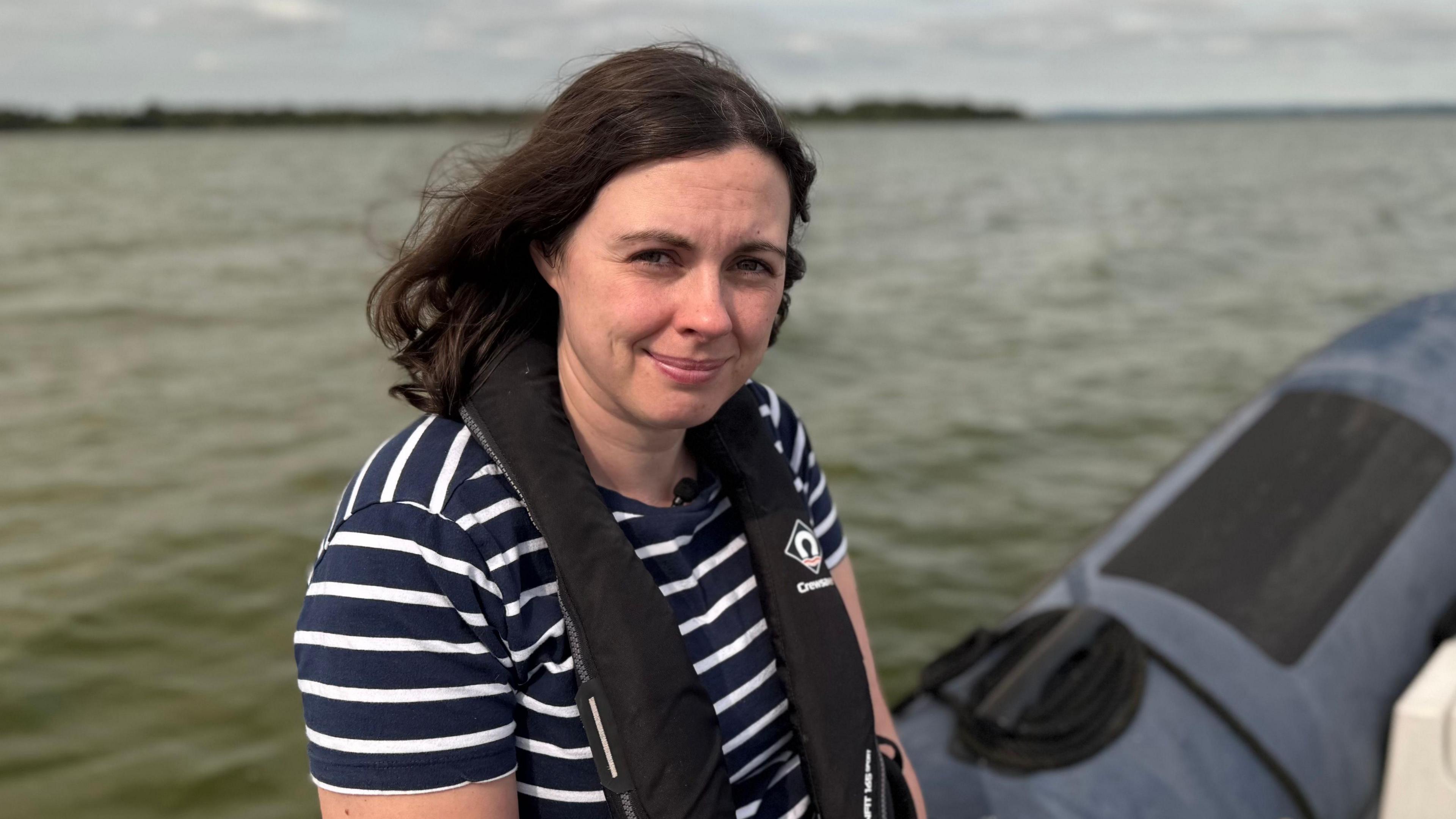 Ciara Laverty is a younger woman with shoulder-length brown hair. She is wearing a navy and white stripe top and a black lifejacket. She is sat on the edge of a boat that is on Lough Neagh but the background is blurred.