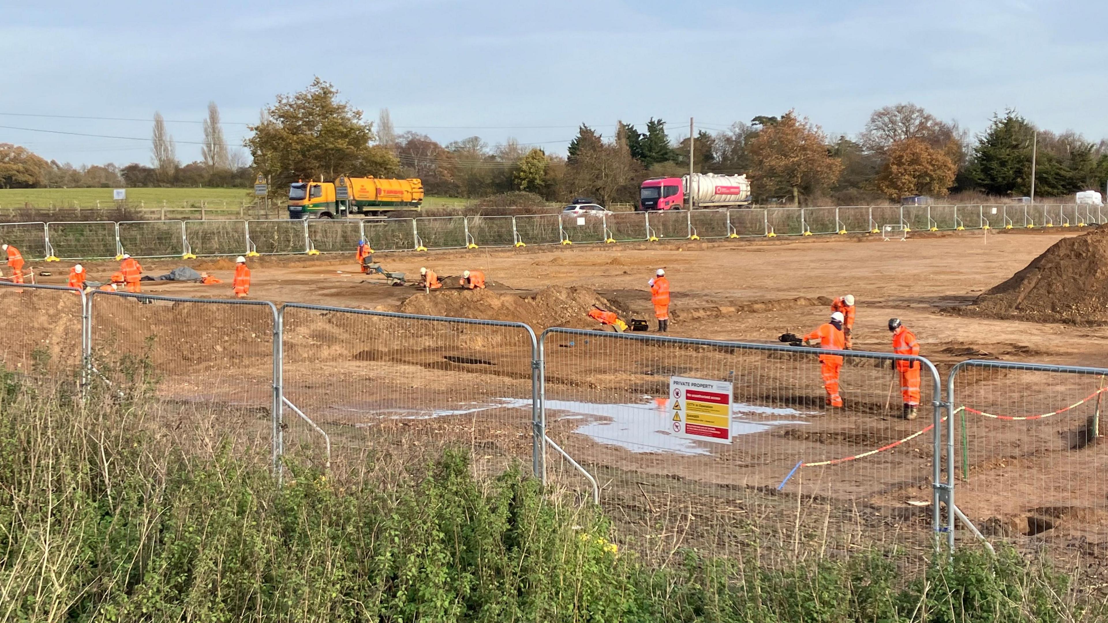 Archaeologists wearing hats and orange fluorescent jackets and trousers are working in an industrial landscape of bare sandy soil surrounded by a metal fence   