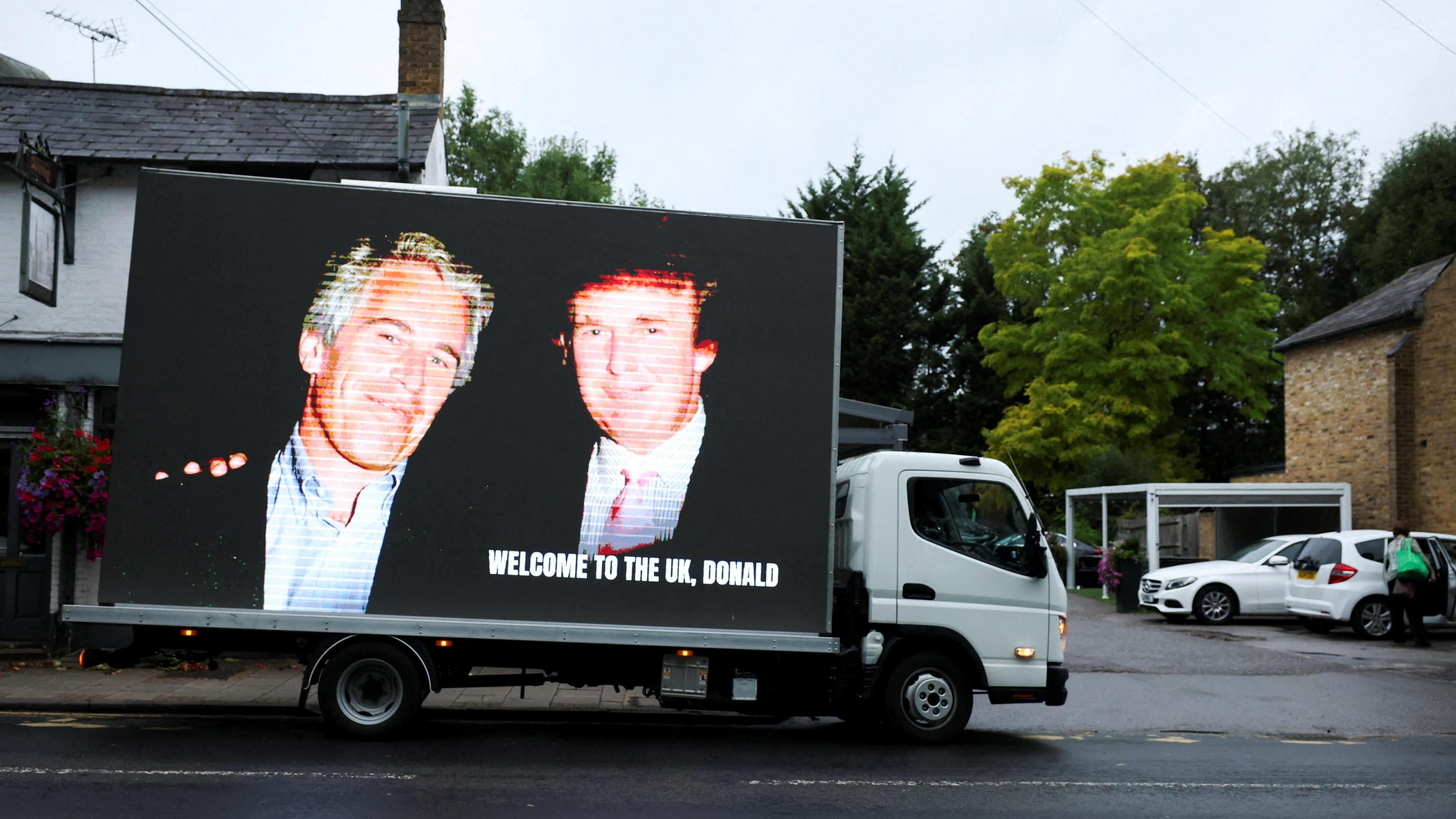 A van with a large screen on its side is parked outside a building in Windsor. The screen displays an image of Trump and Epstein. The two men are wearing suits in and posing together, smiling against a black background. A message in the bottom right of the image reads in all capitals: "Welcome to the UK, Donald." The van is pictured in a small car park with a few white cars parked at the side, under an overcast sky.