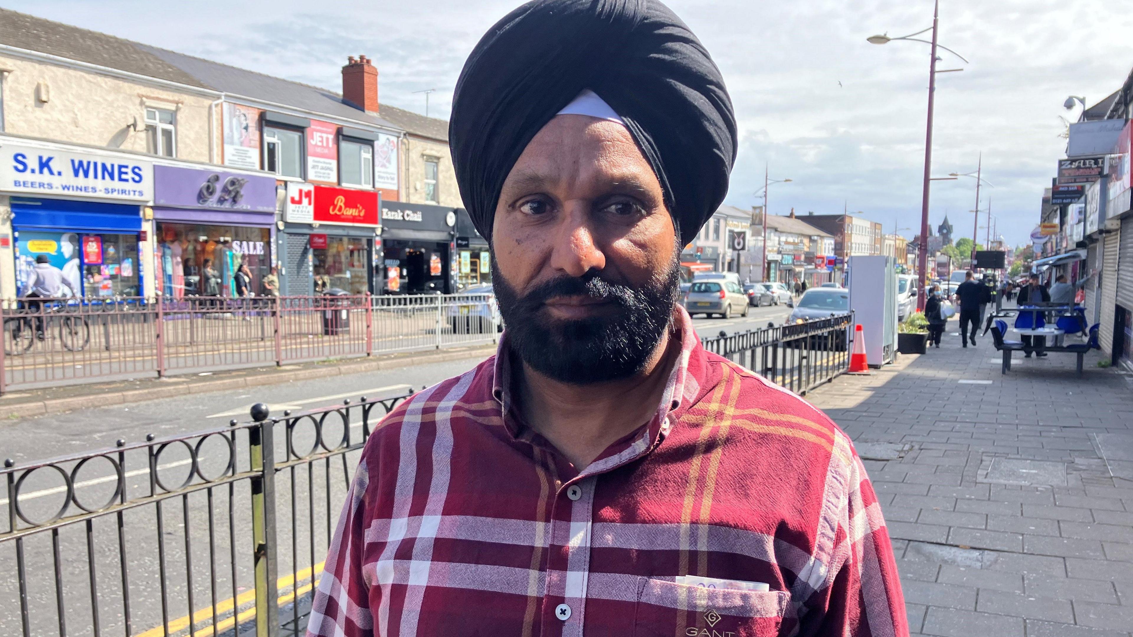 Paramjit Singh Behal looking at the camera. He is stood on the street with shops and railings behind him and is wearing a check red shirt.  