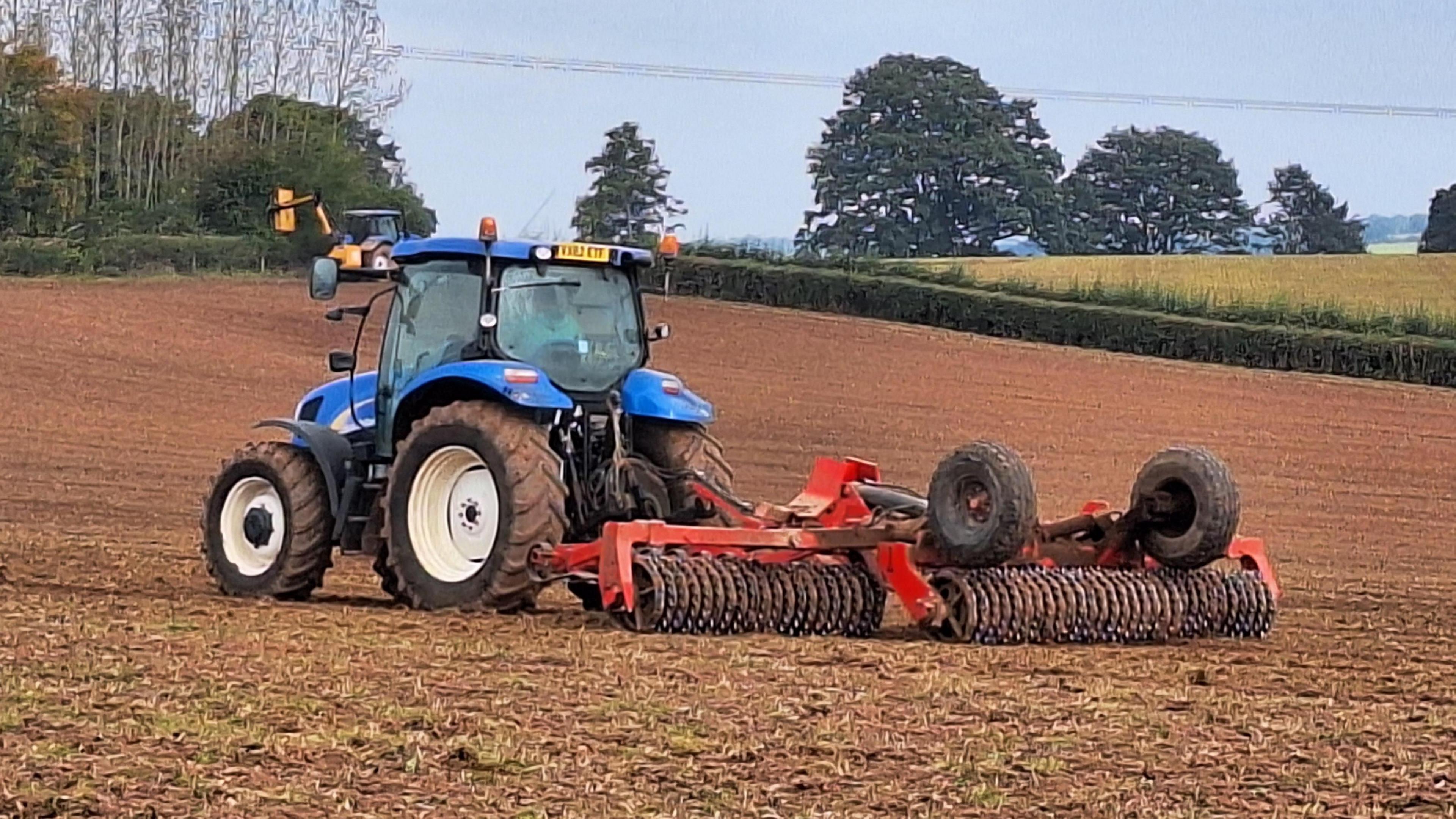 A tractor pulling machinery on a field in Bodenham Herefordshire with trees in the background.