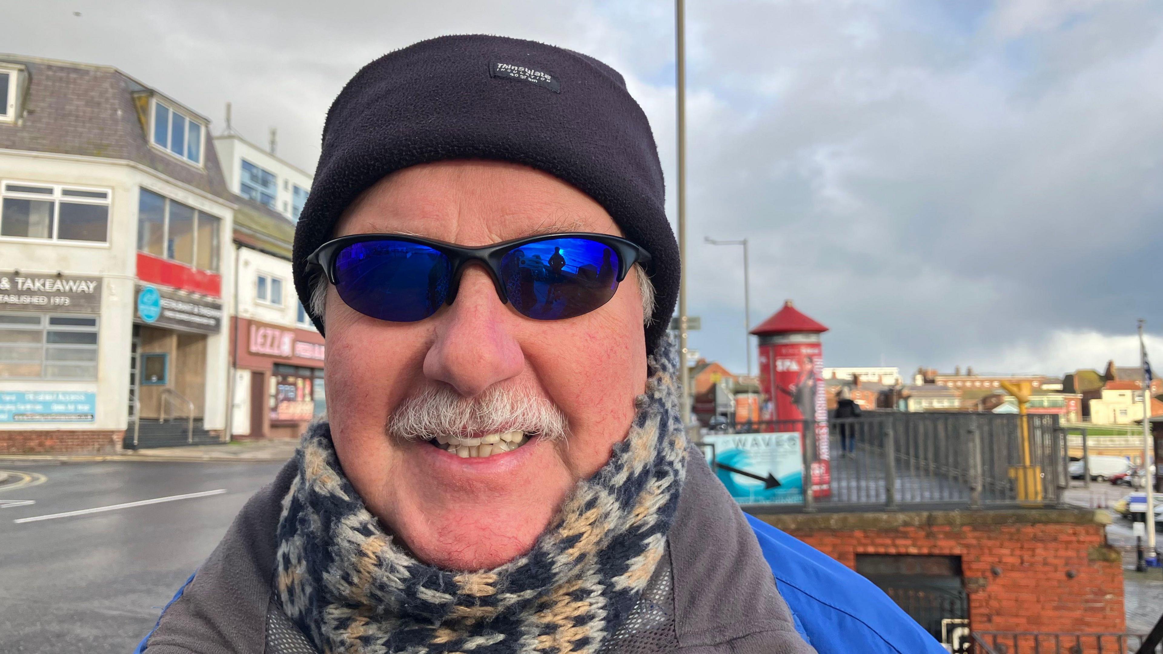 A man with blue sunglasses and a black beanie is smiling into the camera. He has a grey moustache and is wearing a yellow, black and white scarf and coat. Behind him is a road with various shops.