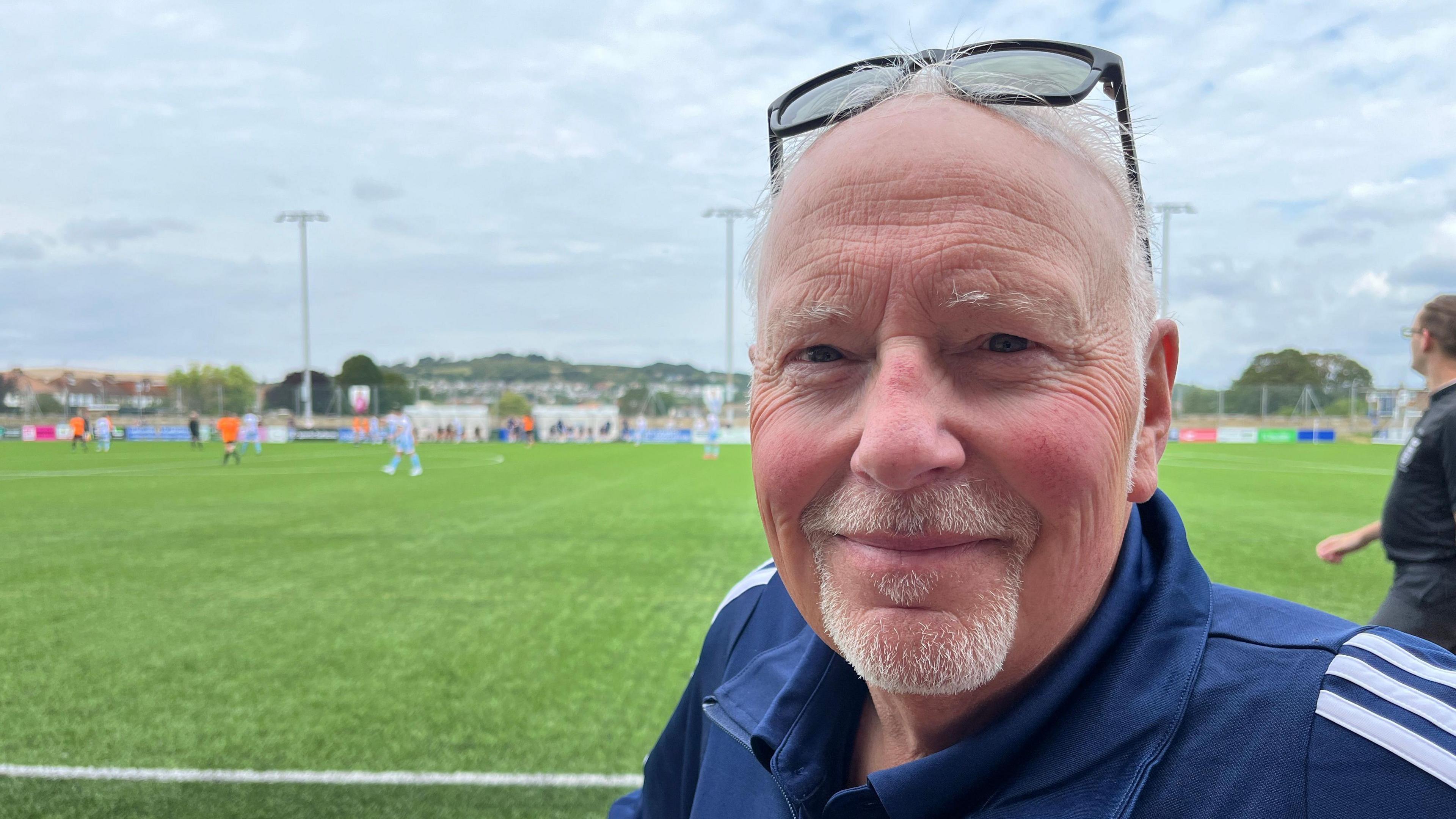 A man wearing a navy blue tracksuit top stood in front of a football pitch. He has sunglasses perched on the top of his head. He has wispy white hair, cut short, and a small beard and moustache.