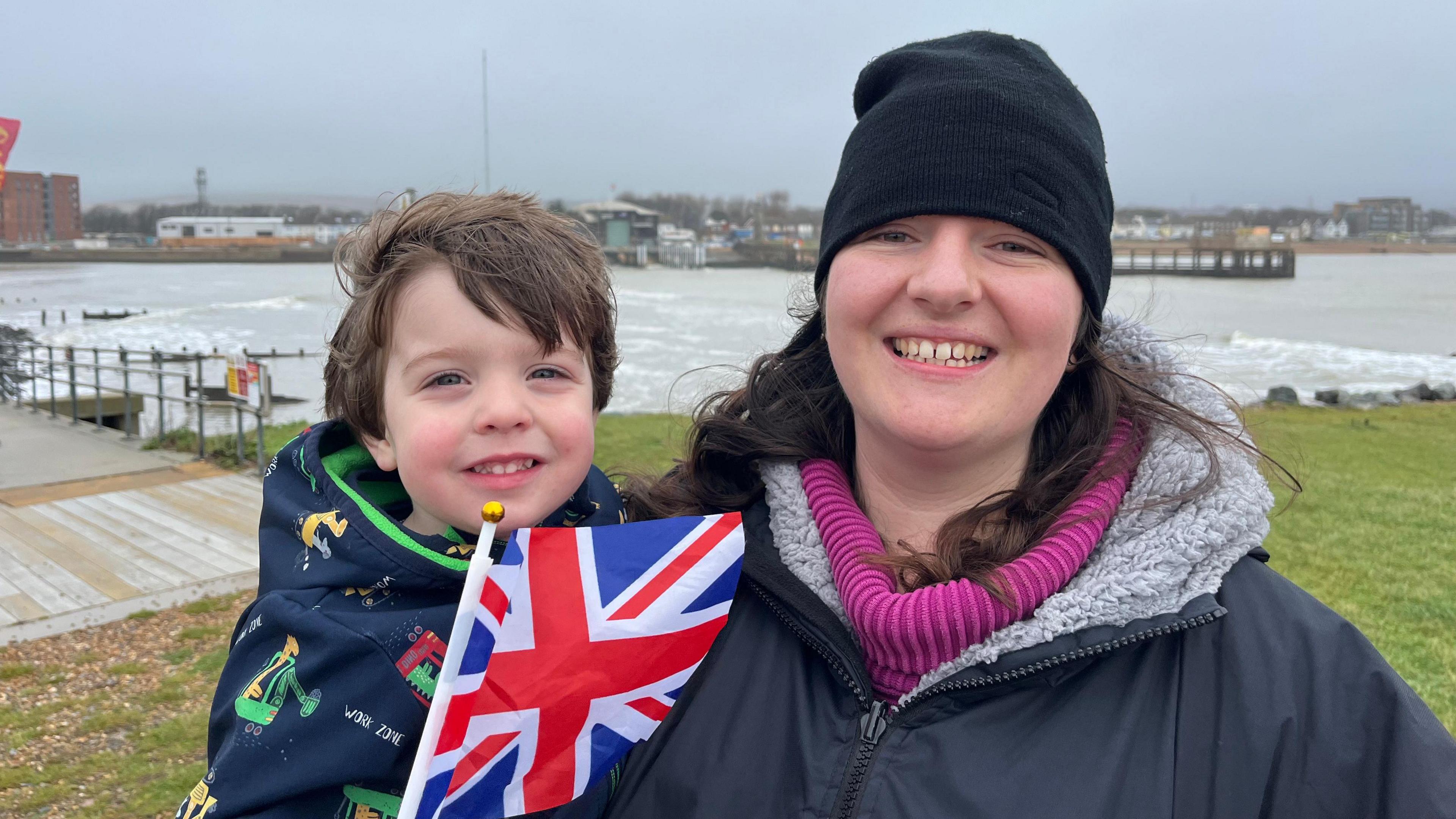 A young boy with brown hair waving a Union Flag next to a woman with long brown hair wearing a blue coat and pink jumper with black hat. They are standing on grass above the sea. In the background are various brown buildings