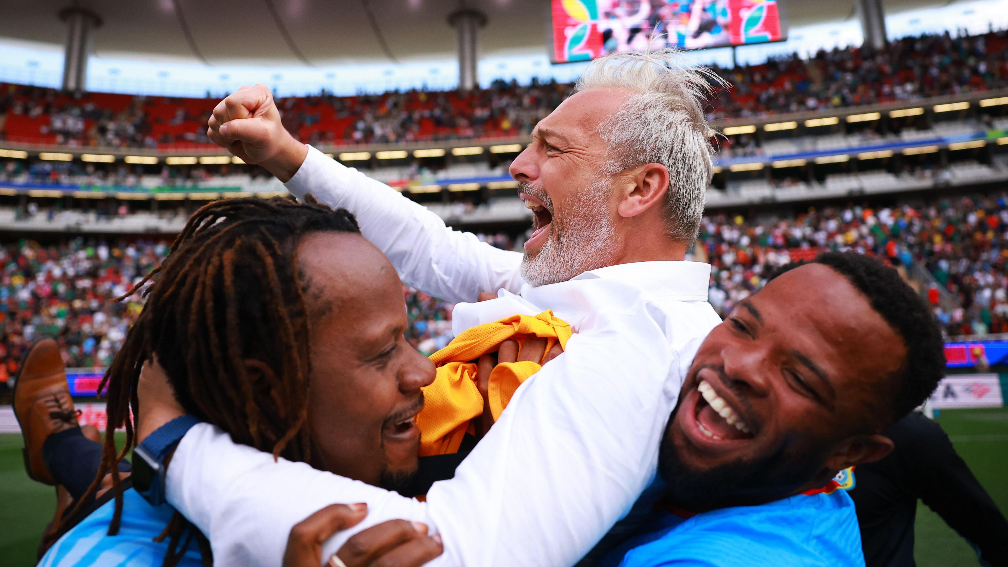 DR Congo coach Sebastien Desabre is seen in a long-sleeved smart white shirt being lifted up by two players, with one grabbing him from behind and the other from in front. Desabre is punching the air and screaming with joy as he celebrates his side's World Cup qualification. In the background can be seen the high tiers of a stand with red seats