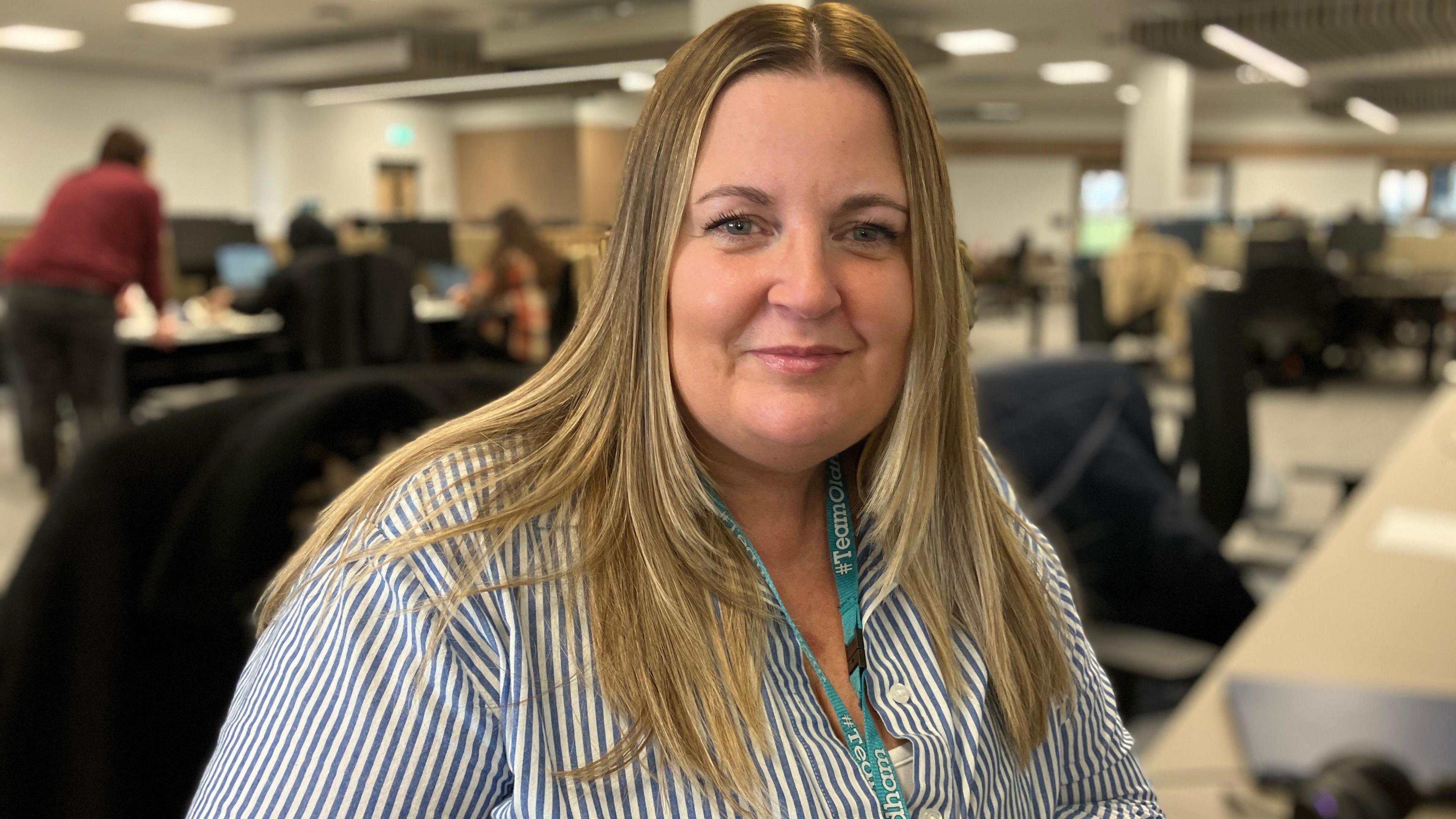 Social worker Tracy Hynes sits in an office wearing a blue and white striped shirt and a teal lanyard. She has mid length brown-blonde hair.