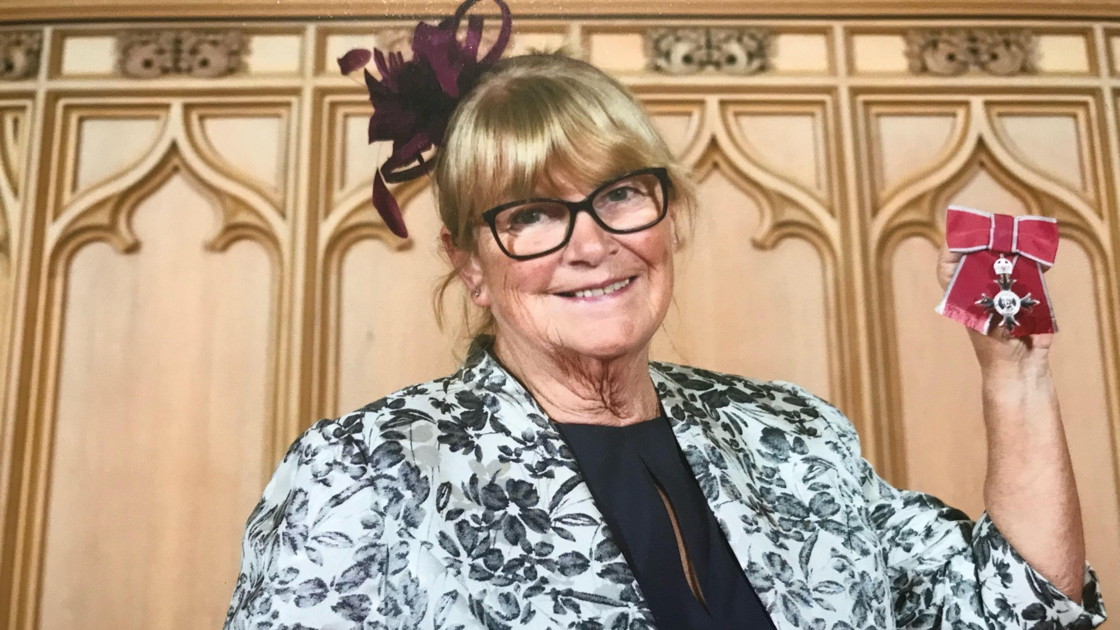 A woman with a blonde fringe is wearing a black top and a white floral jacket. She is holding up her MBE medal with its red ribbon.